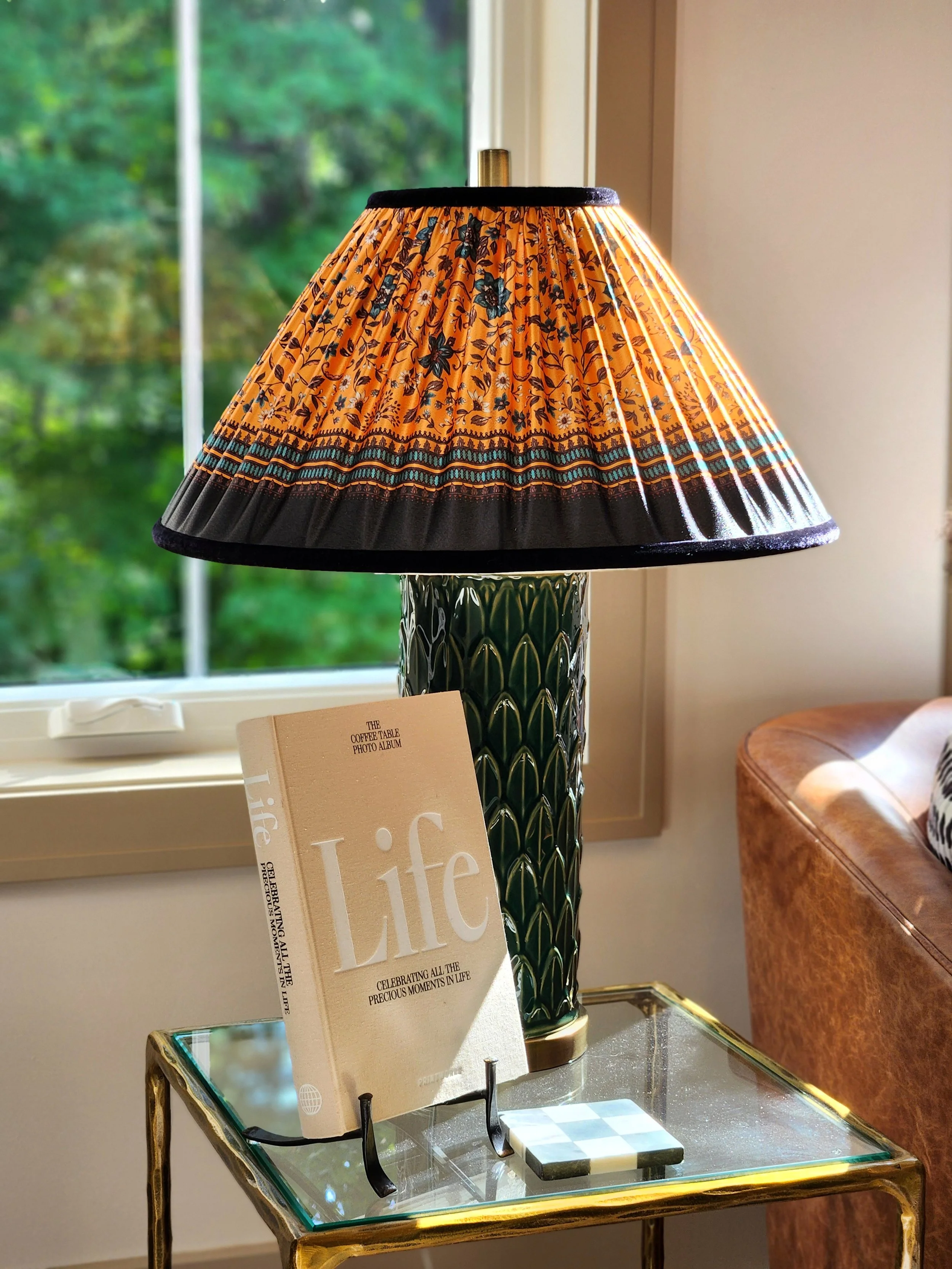 Decorative table lamp with a floral-patterned lampshade, a book titled 'Life' on a glass table, and a small object on the table, with a window showing green trees outside.