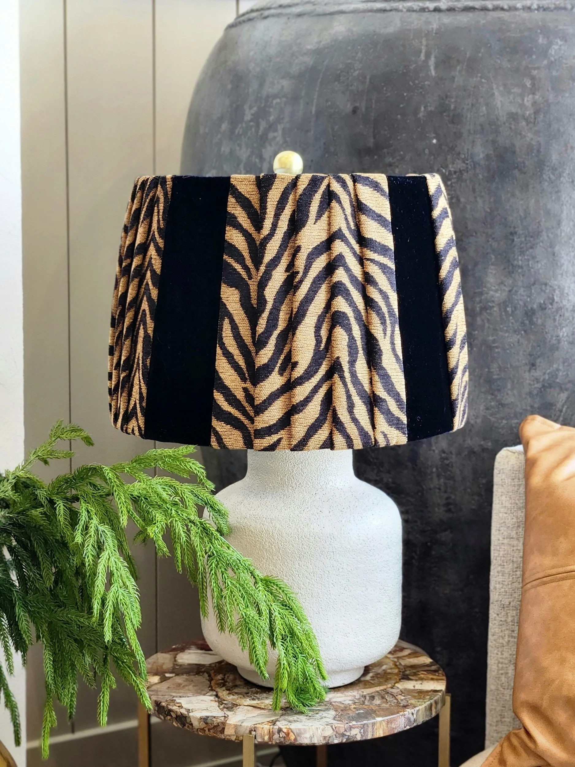 Decorative lamp with zebra-patterned shade on a textured table, next to green foliage and a leather cushion.
