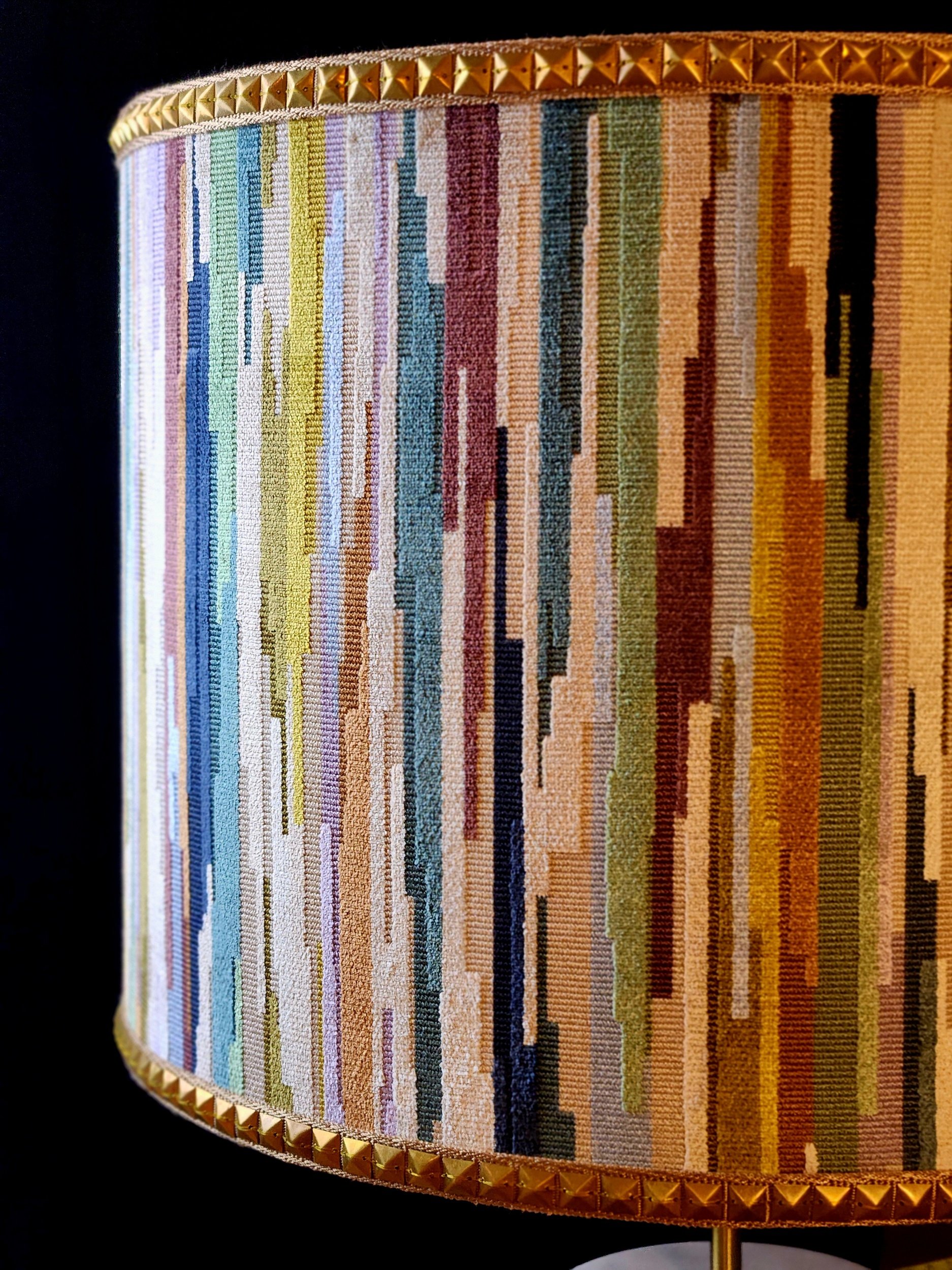 Close-up of a cylindrical, colorful, pixelated patterned lampshade with gold studded trim at the top and bottom edges, against a dark background.