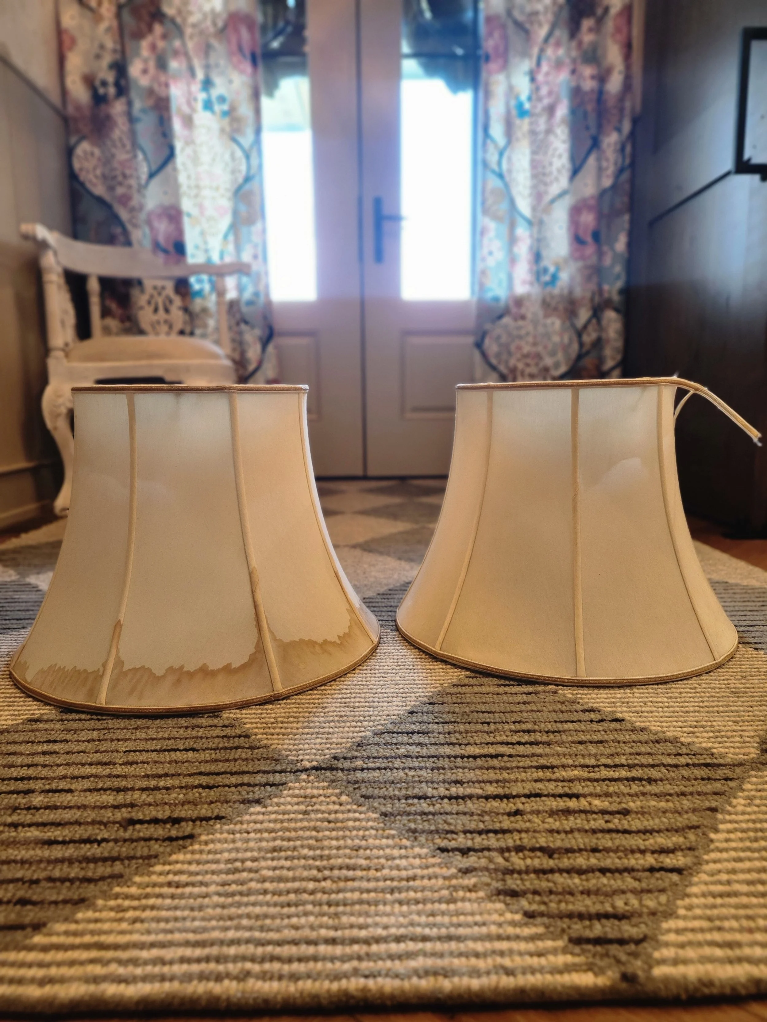 Two broken lampshades on a patterned rug in front of a door with floral curtains.