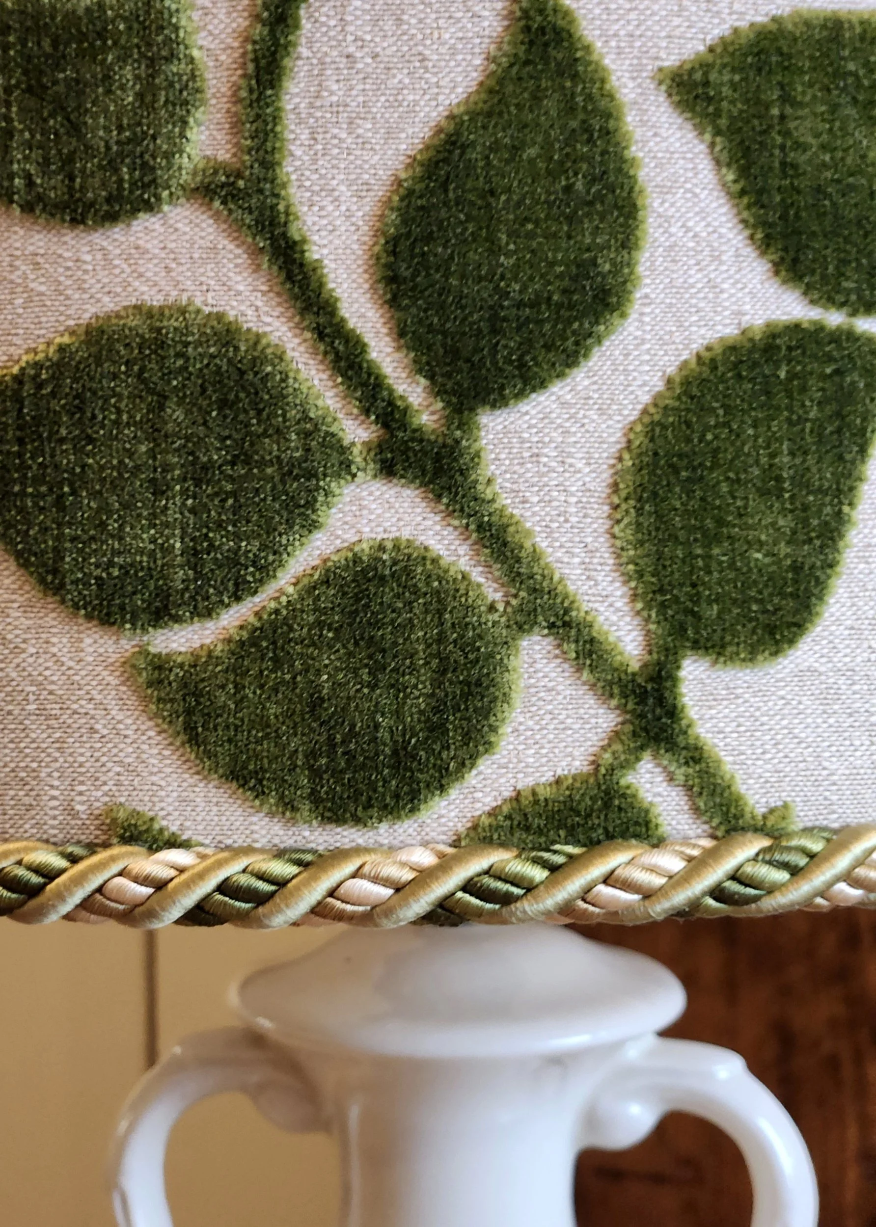 Close-up of a decorative fabric with green velvet leaves and a braided trim, placed on top of a small white ceramic vase.