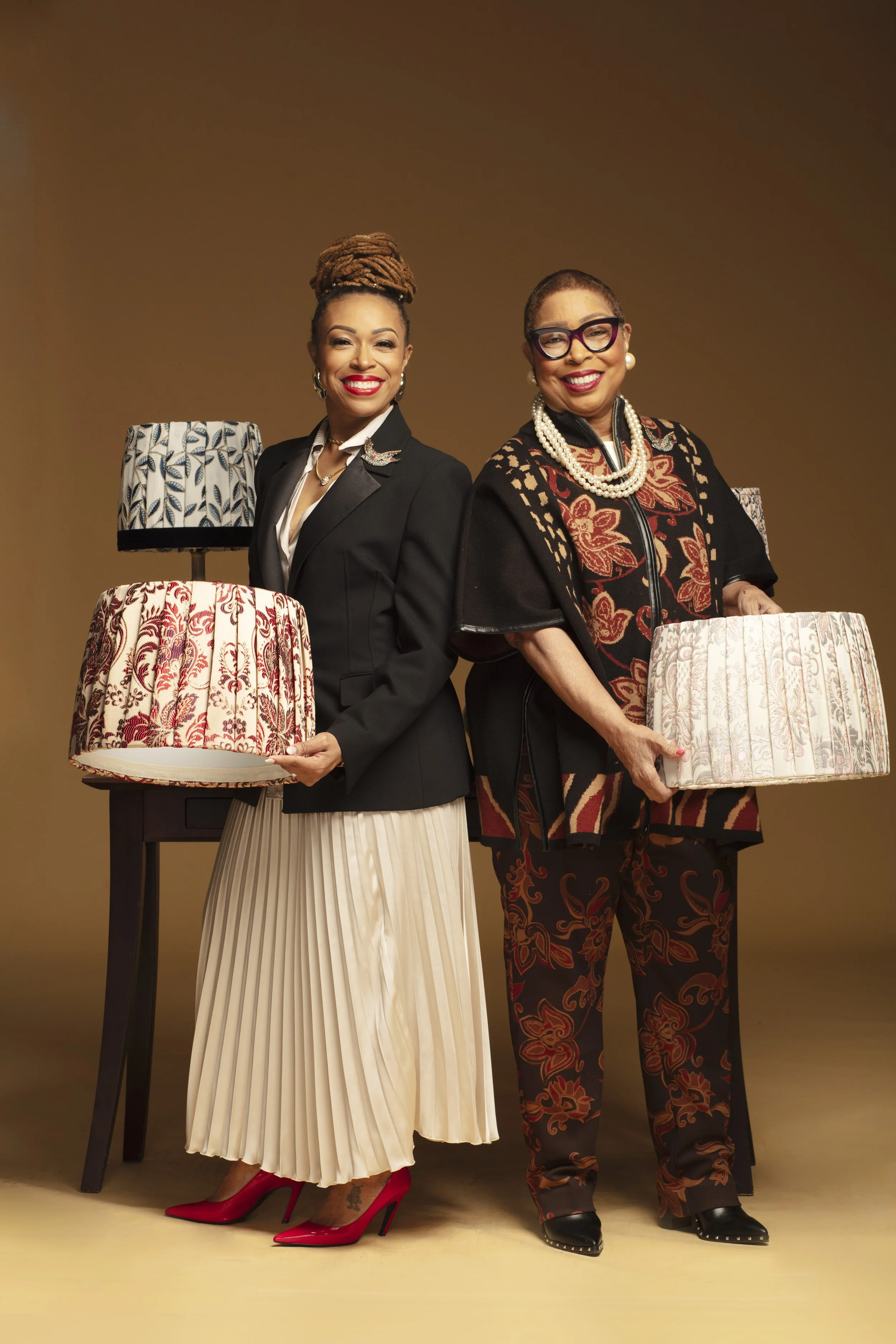 Two women standing with fabric-covered lampshades, wearing stylish outfits against a plain brown background.