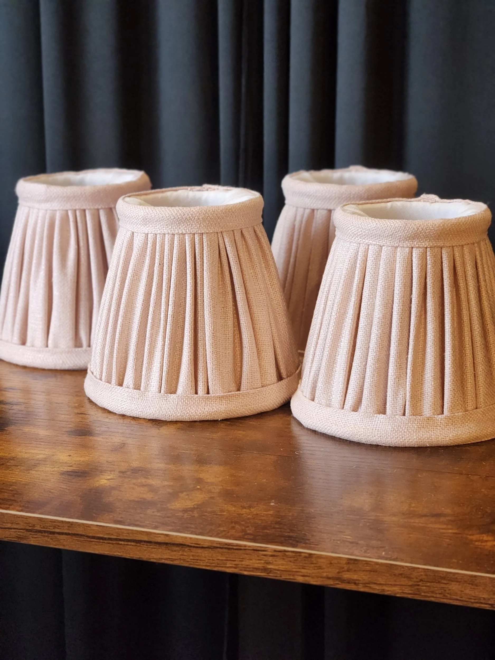 Four blush pink pleated lampshades on a wooden surface with a black curtain background.
