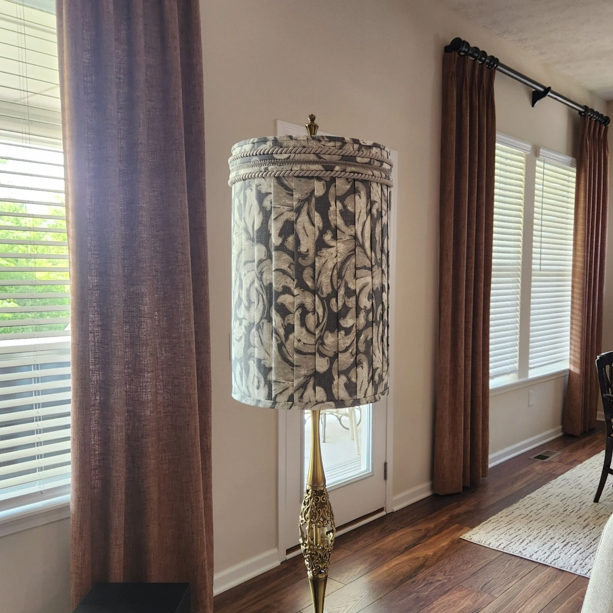 Decorative floor lamp with ornate base and patterned lampshade in a living room with wooden floor and curtains.