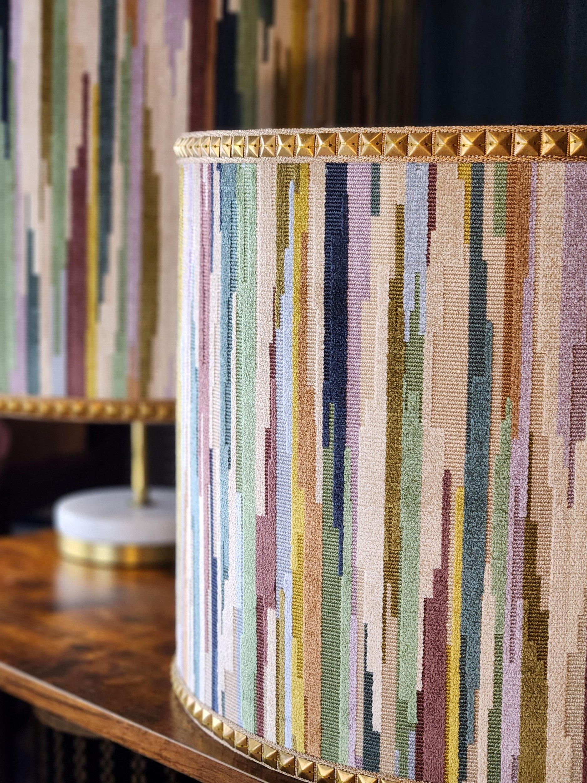 Close-up of a fabric-covered lampshade with multicolored vertical stripes and gold pyramid-shaped studs along the top and bottom edges, placed on a wooden table with a blurred bookshelf in the background.