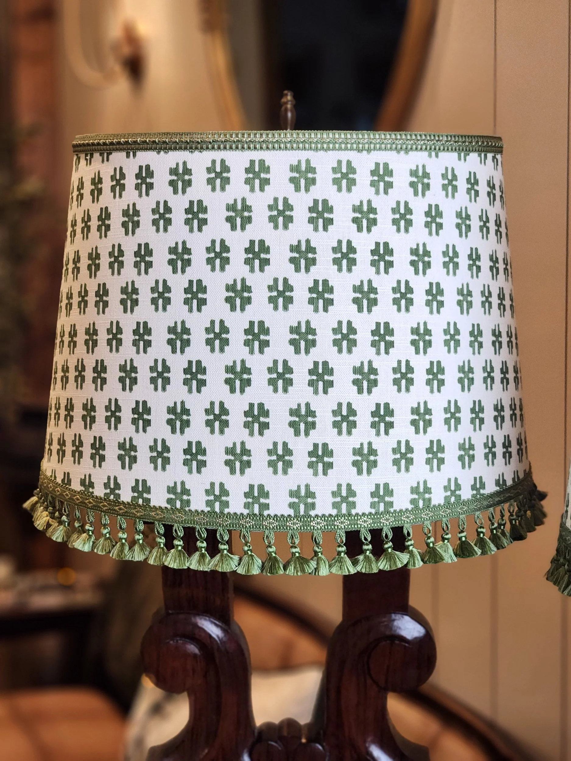 A decorative lampshade with a repeating pattern of green four-leaf clovers and green fringe trim, placed on a wooden lamp base.