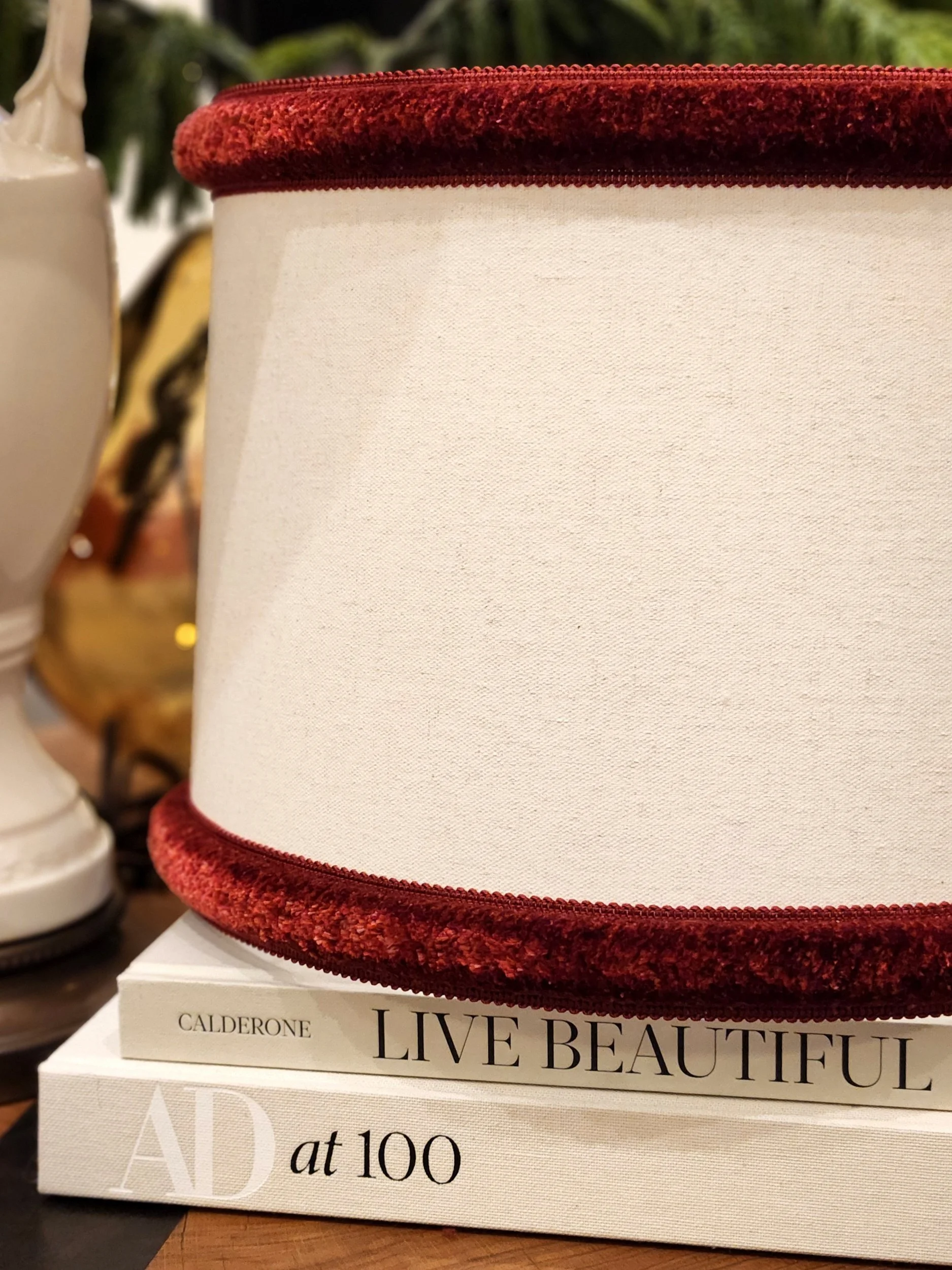 A cream-colored fabric lampshade with red velvet trim, sitting on top of two white books titled 'LIVE BEAUTIFUL' and 'AD at 100'.