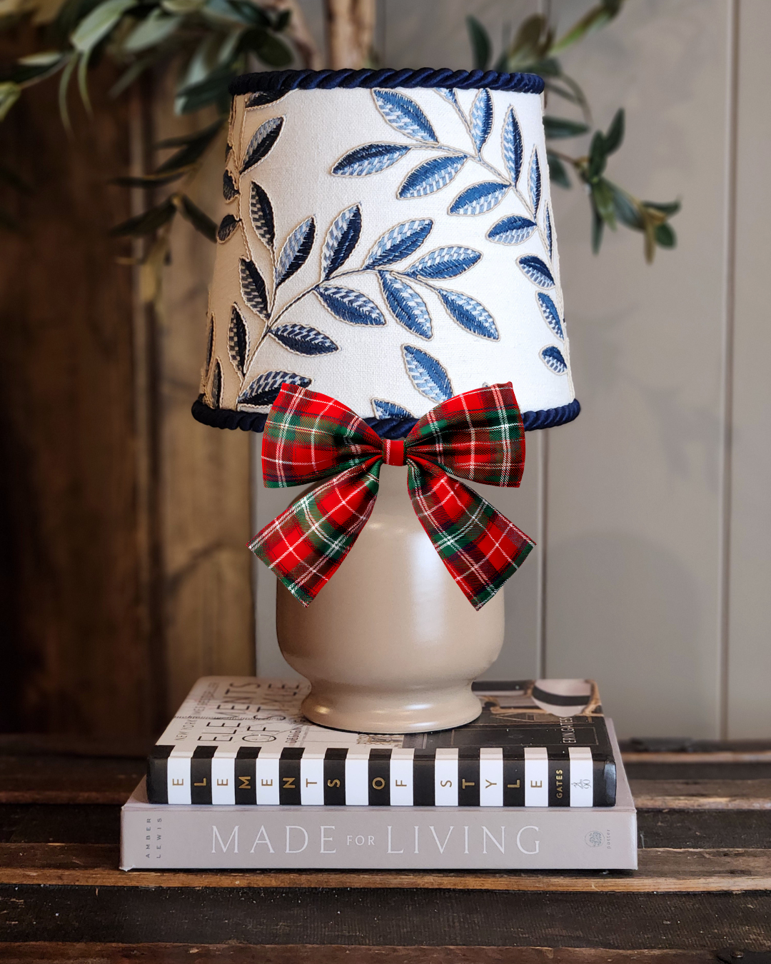 A decorative table lamp with a white base, a lampshade featuring blue embroidered leaves, and a red and green plaid bow, placed on a stack of books on a wooden surface.