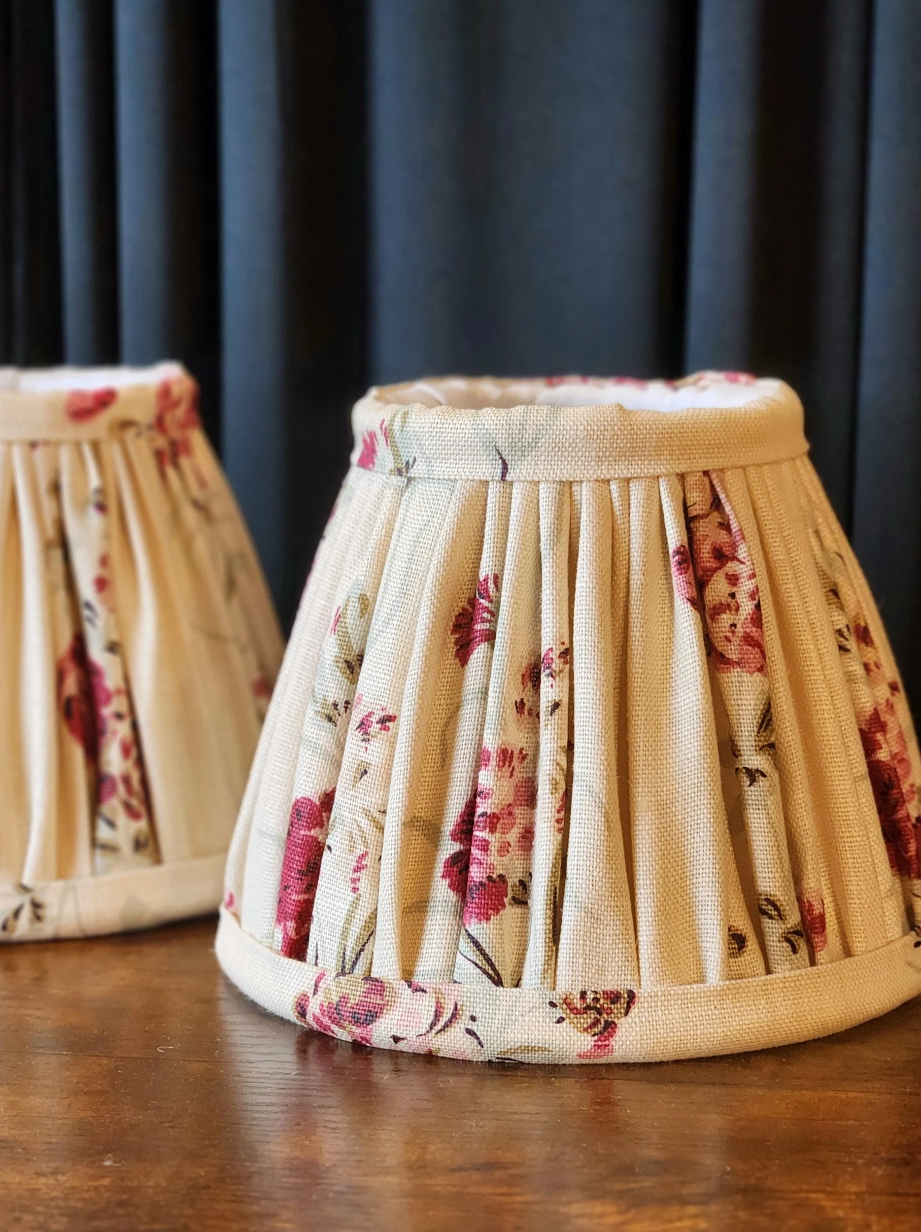 Close-up of a fabric lampshade with floral patterns, sitting on a wooden surface, against a dark curtain background.