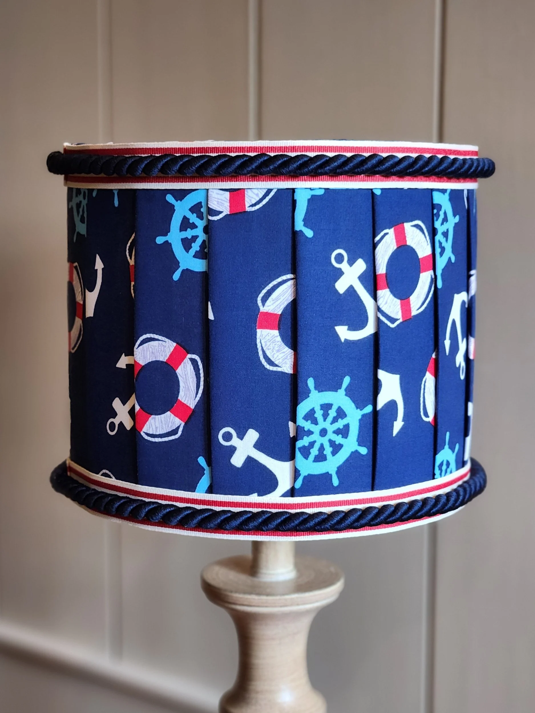 A cylindrical lampshade with a nautical themed fabric featuring anchors, ship wheels, and life preservers, with navy blue, white, and red colors, held by a wooden lamp base.