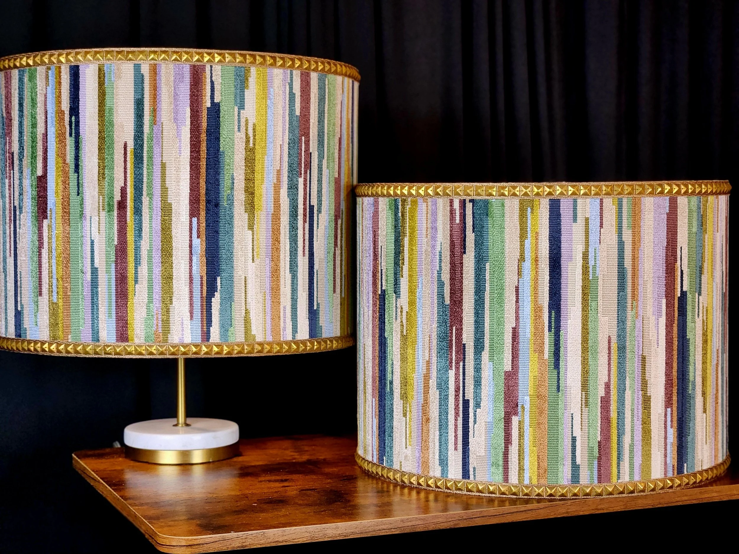A table lamp with a colorful, vertically striped fabric shade and gold trim on top and bottom, sitting on a wooden surface with a dark background.