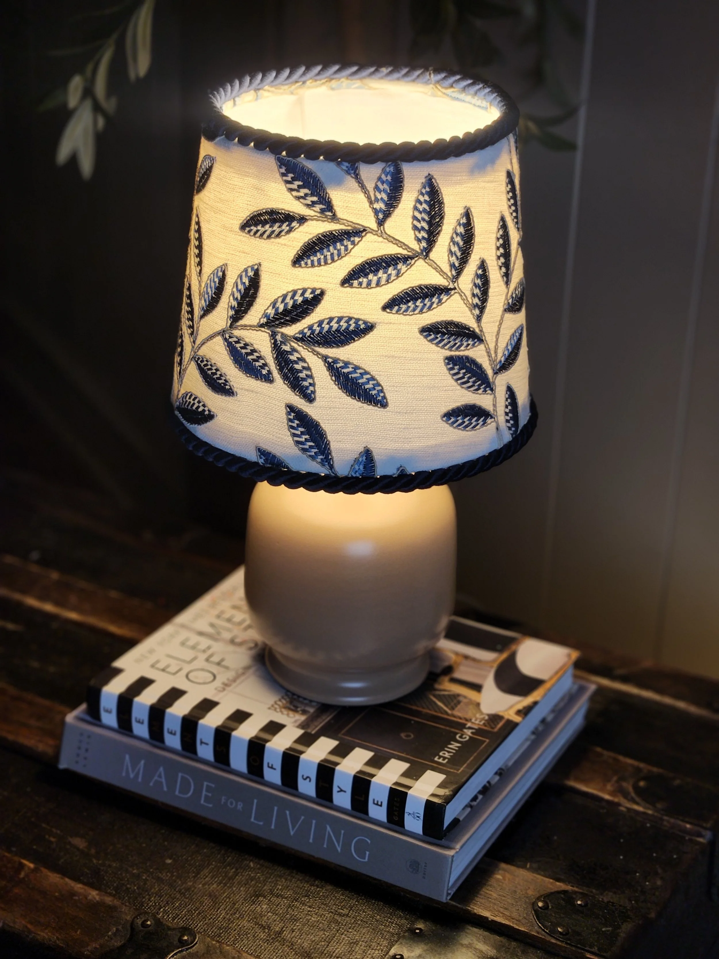 A table lamp with a fabric shade featuring blue and white leaf patterns, placed on two stacked books on a wooden surface.