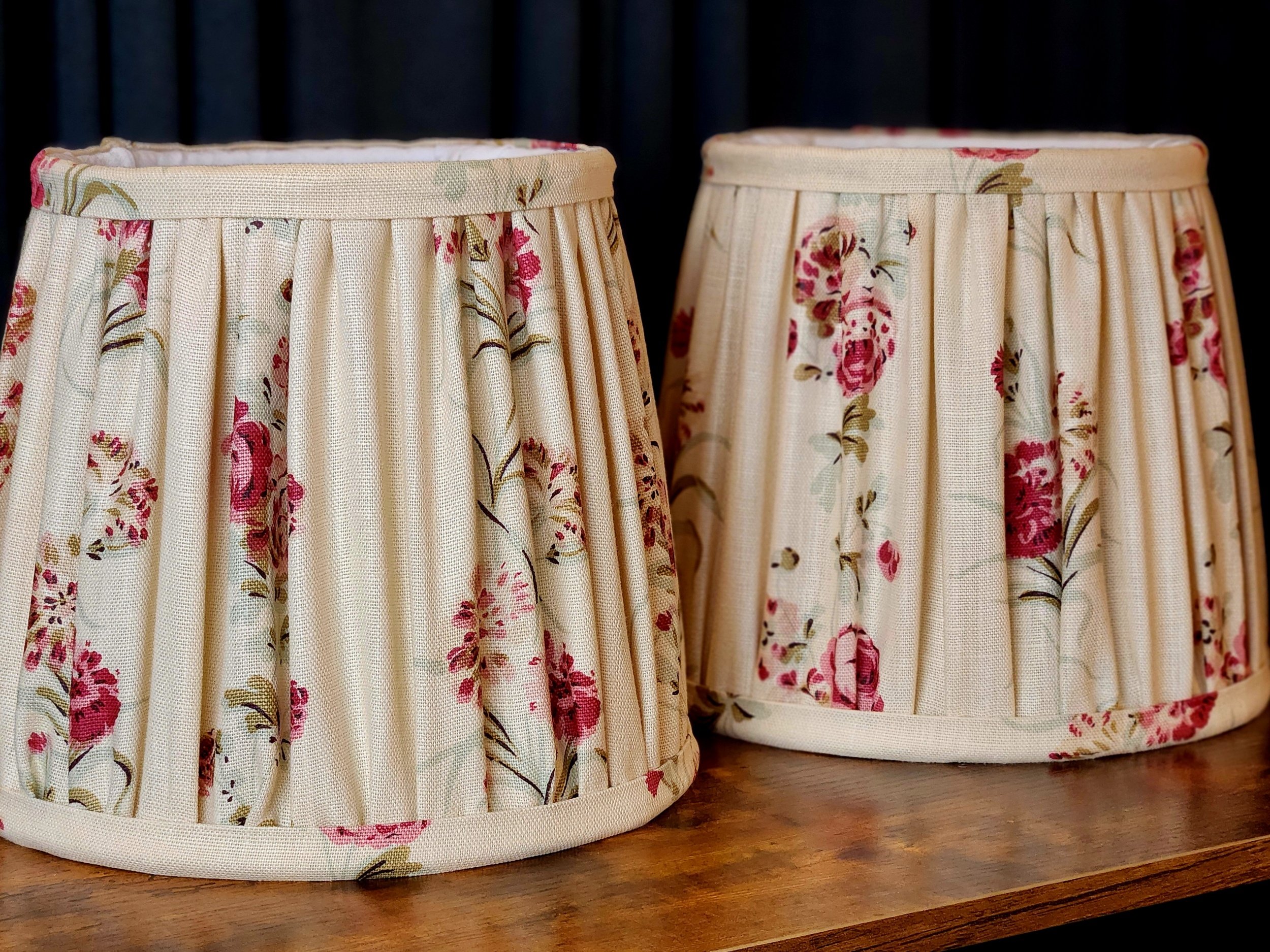 Two fabric lampshades with floral pattern and pleated design, placed on a wooden surface against a dark background.