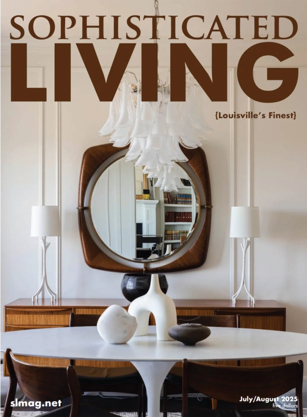 Interior of a sophisticated living room with a round white table, decorative vases, a wooden sideboard, an oval mirror, modern white lamps, and a chandelier, showcasing elegant decor.