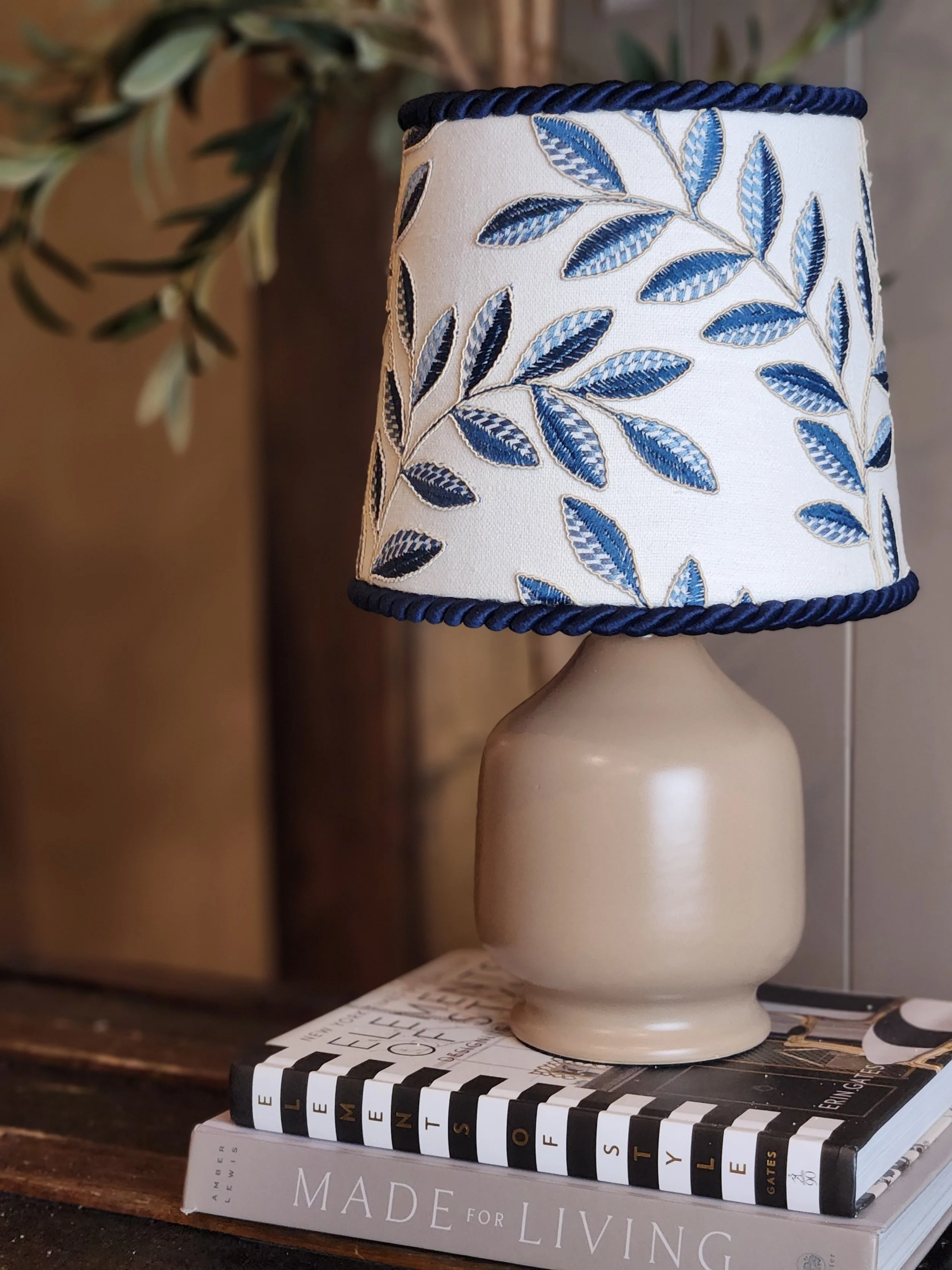 Table lamp with a white lampshade decorated with blue embroidered leaves and a navy braided trim, placed on top of a stack of hardcover books.