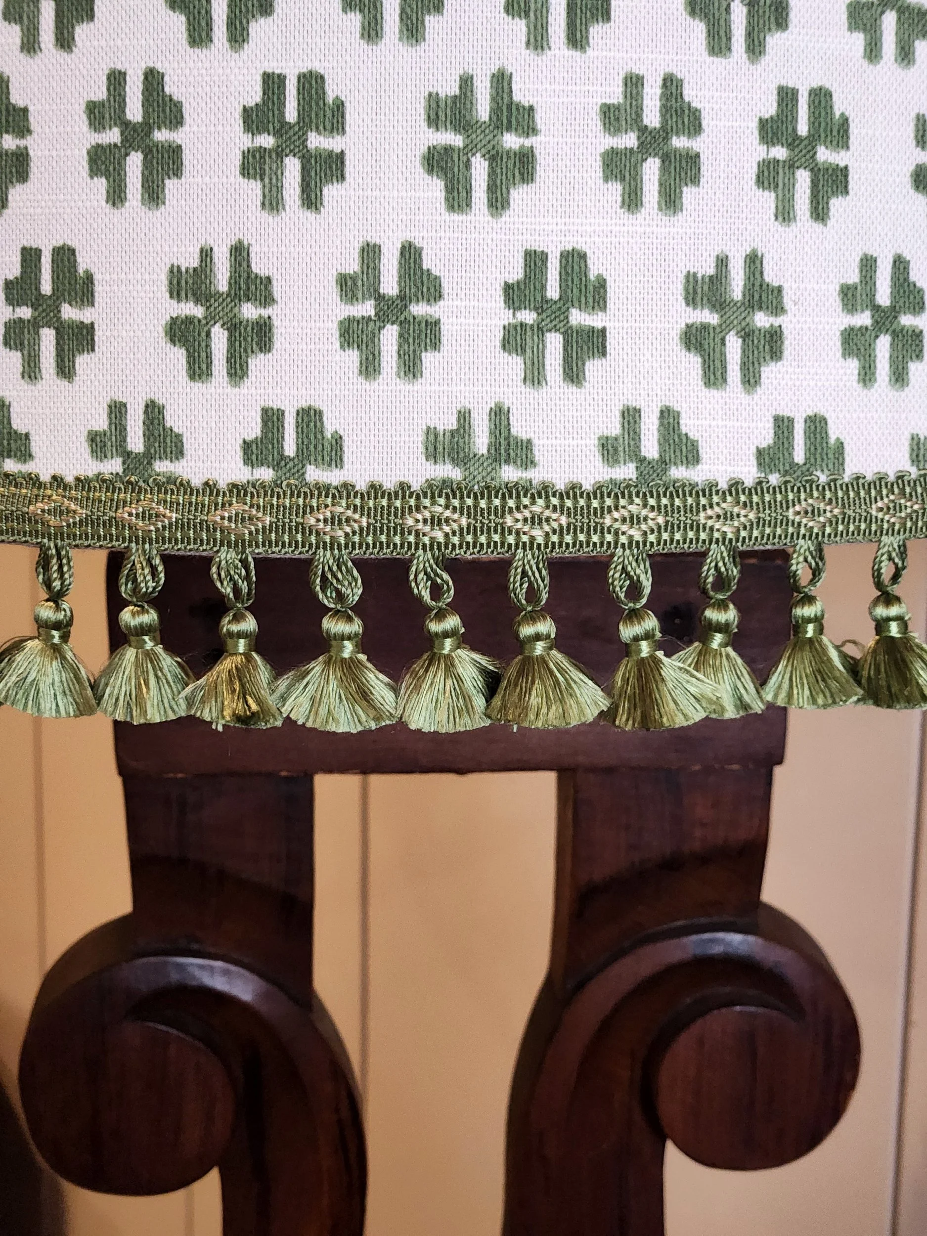 Close-up of a decorative fabric with green clover motifs, green tassels, and trim, draped over a dark wooden furniture piece.