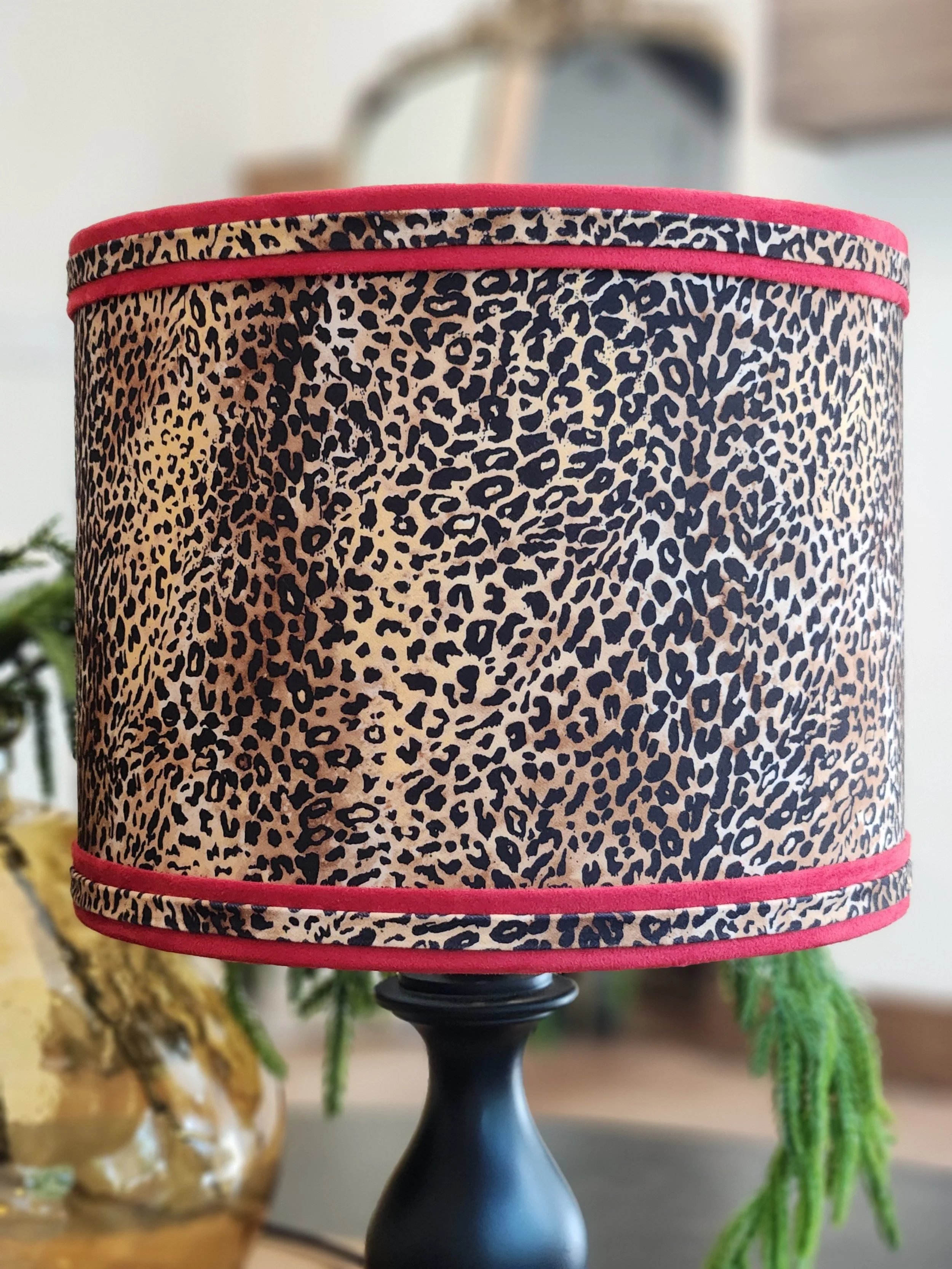 Close-up of a table lamp with a leopard print lampshade and pink trim near the top and bottom, on a table with background plants.