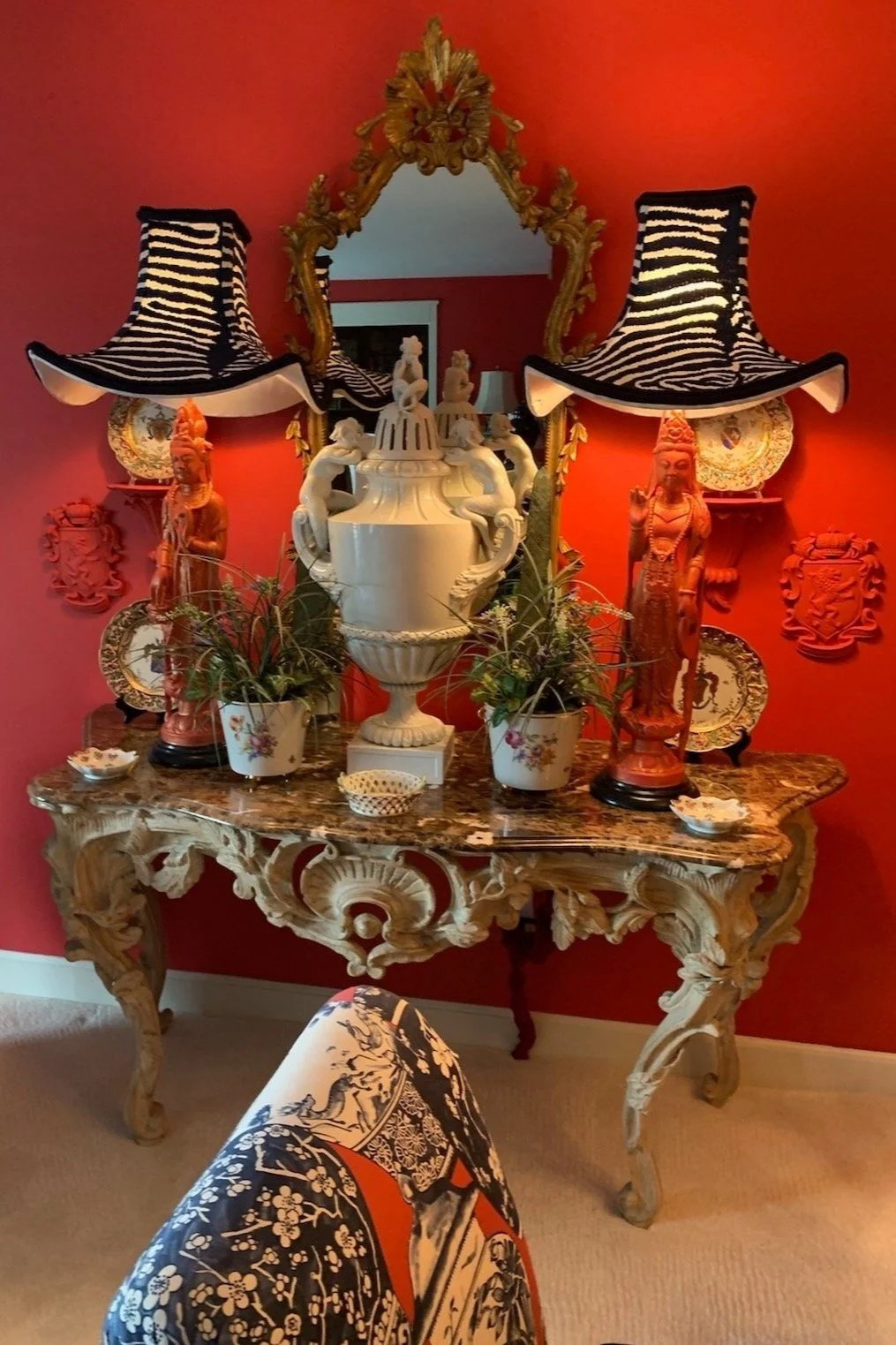 Ornate console table with marble top, red sculptures, striped lamps, decorative mirror, and plants against a red wall.