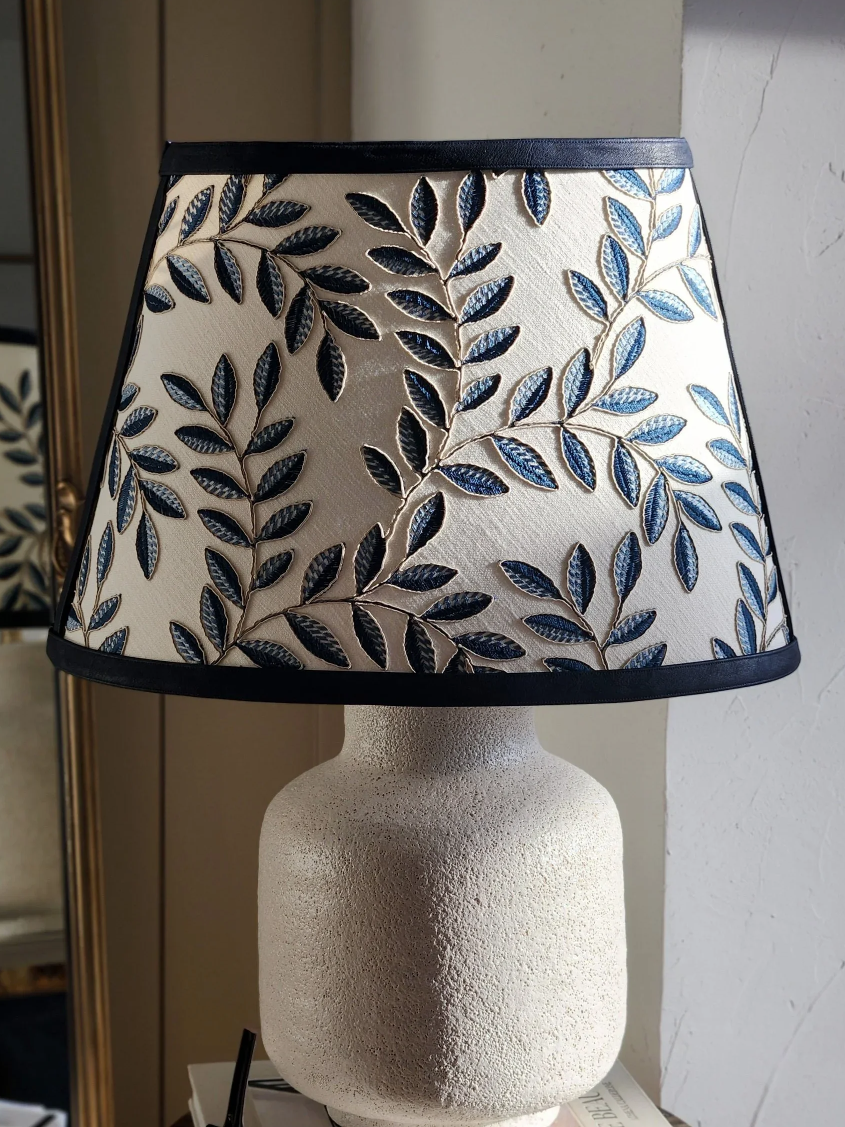 White table lamp with blue and beige leaf-patterned lampshade on a white textured base, placed on a table near a wall.