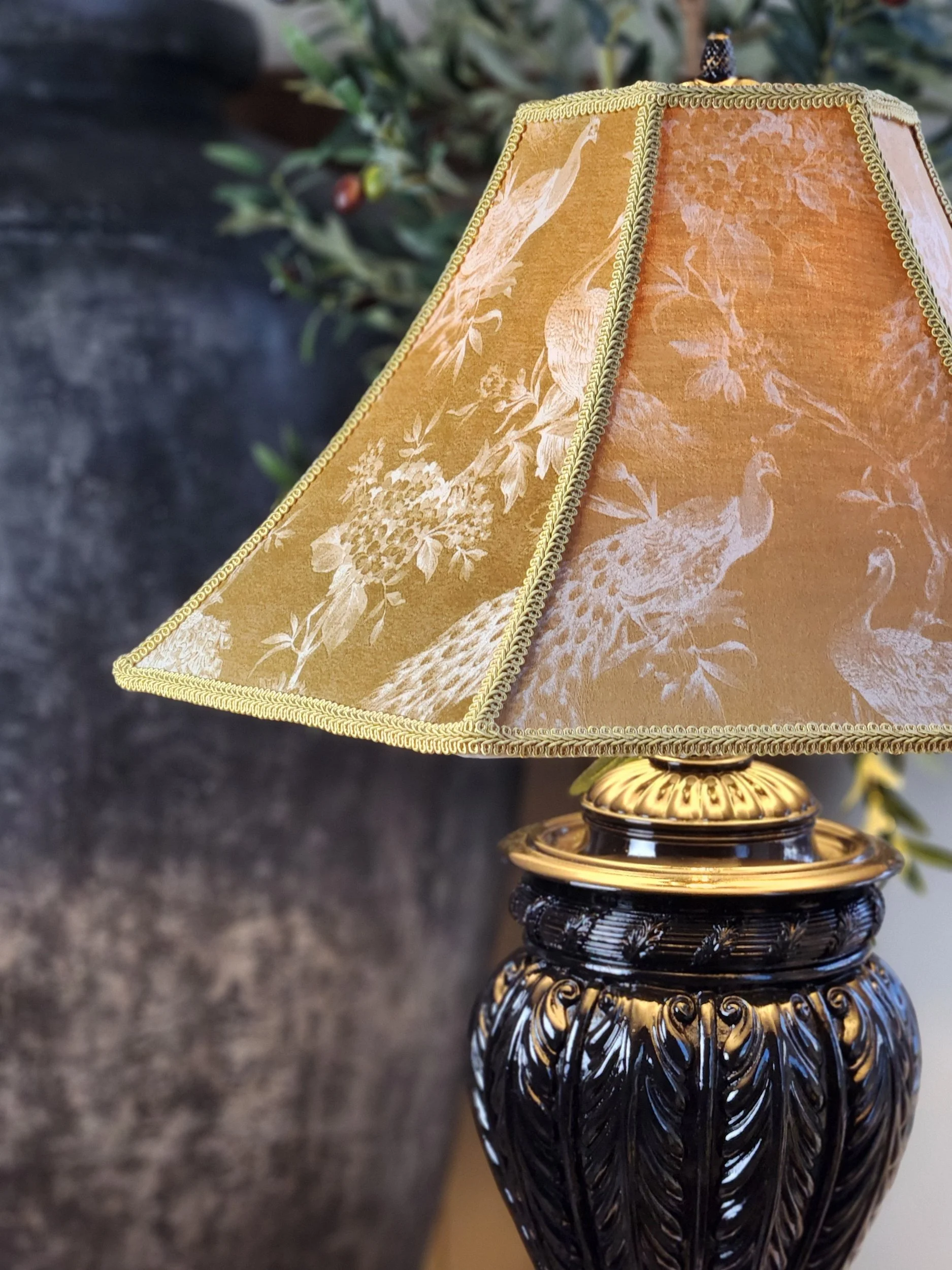 A decorative table lamp with a black and gold base featuring intricate designs and a beige lampshade with peacock and floral patterns.