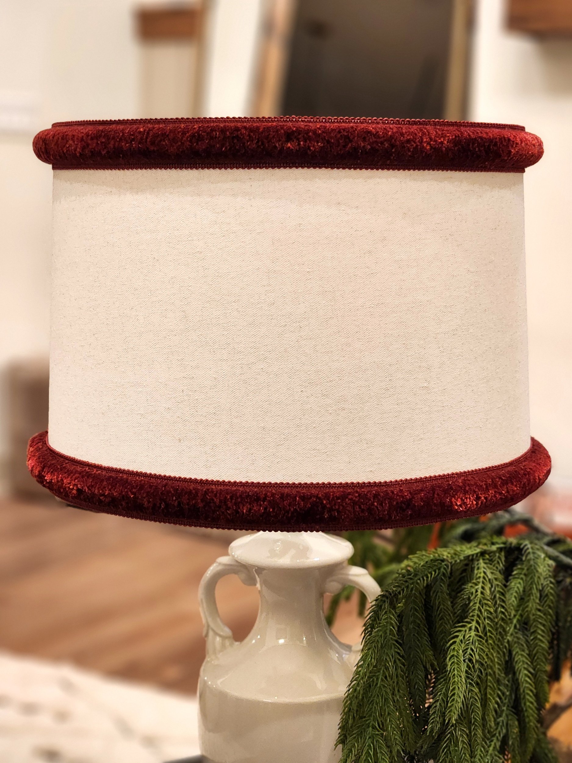Table lamp with a white base and a red velvet lampshade in a room with wooden flooring and blurred furniture.
