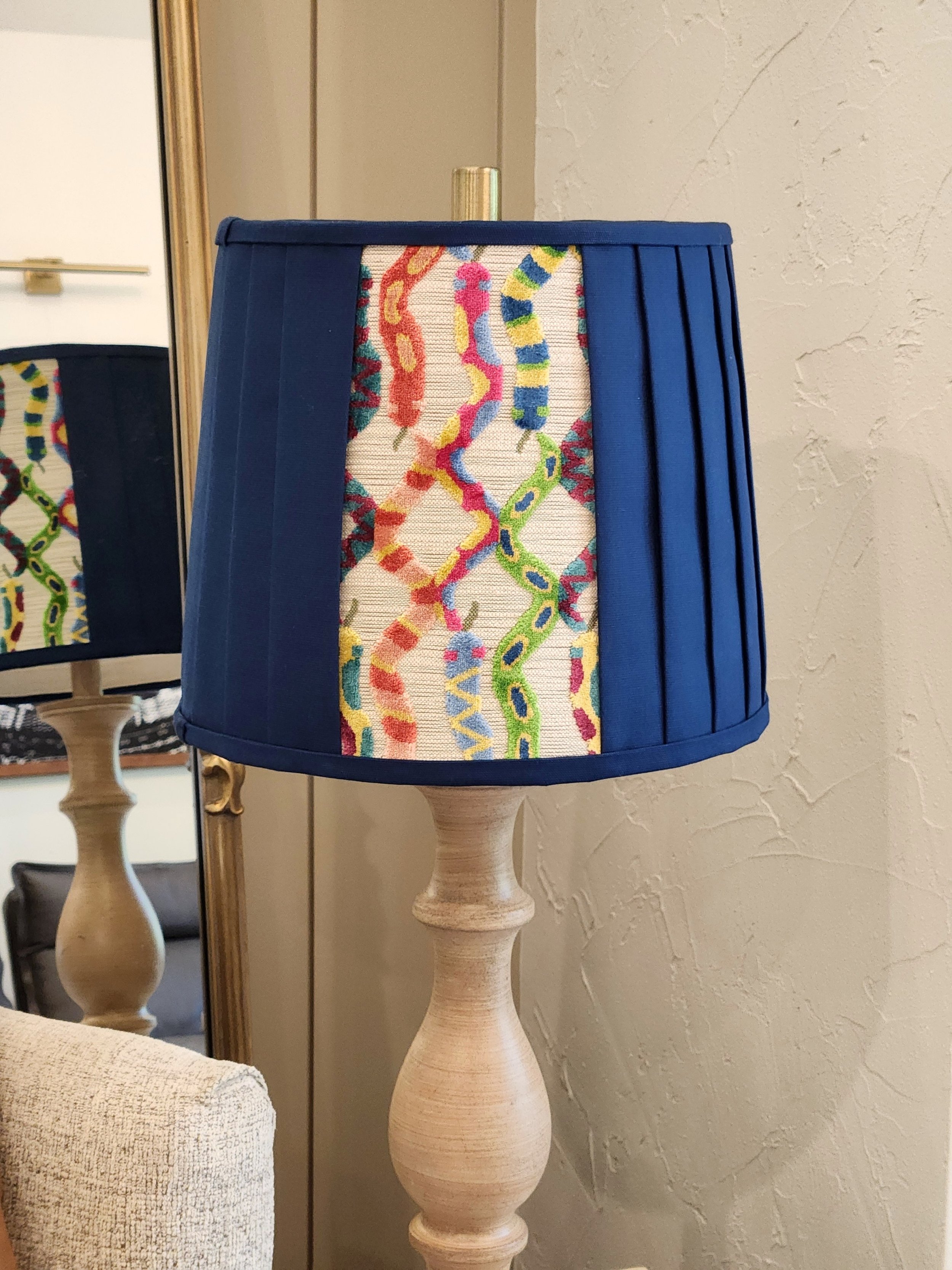 A navy blue lampshade with a striking patterned panel of colorful swirls. The pleated design adds texture, and the lamp sits on a light wooden base, in front of a mirror.