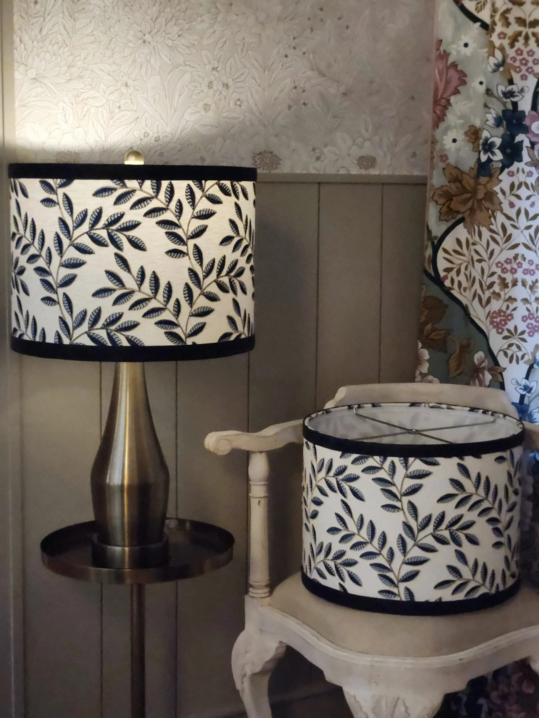 A table lamp and a fabric lampshade with a matching leaf pattern. The lamp has a metallic base and a shade with navy and white leaves. The lampshade, sitting on a vintage white side table, has the same leaf pattern with a navy border.