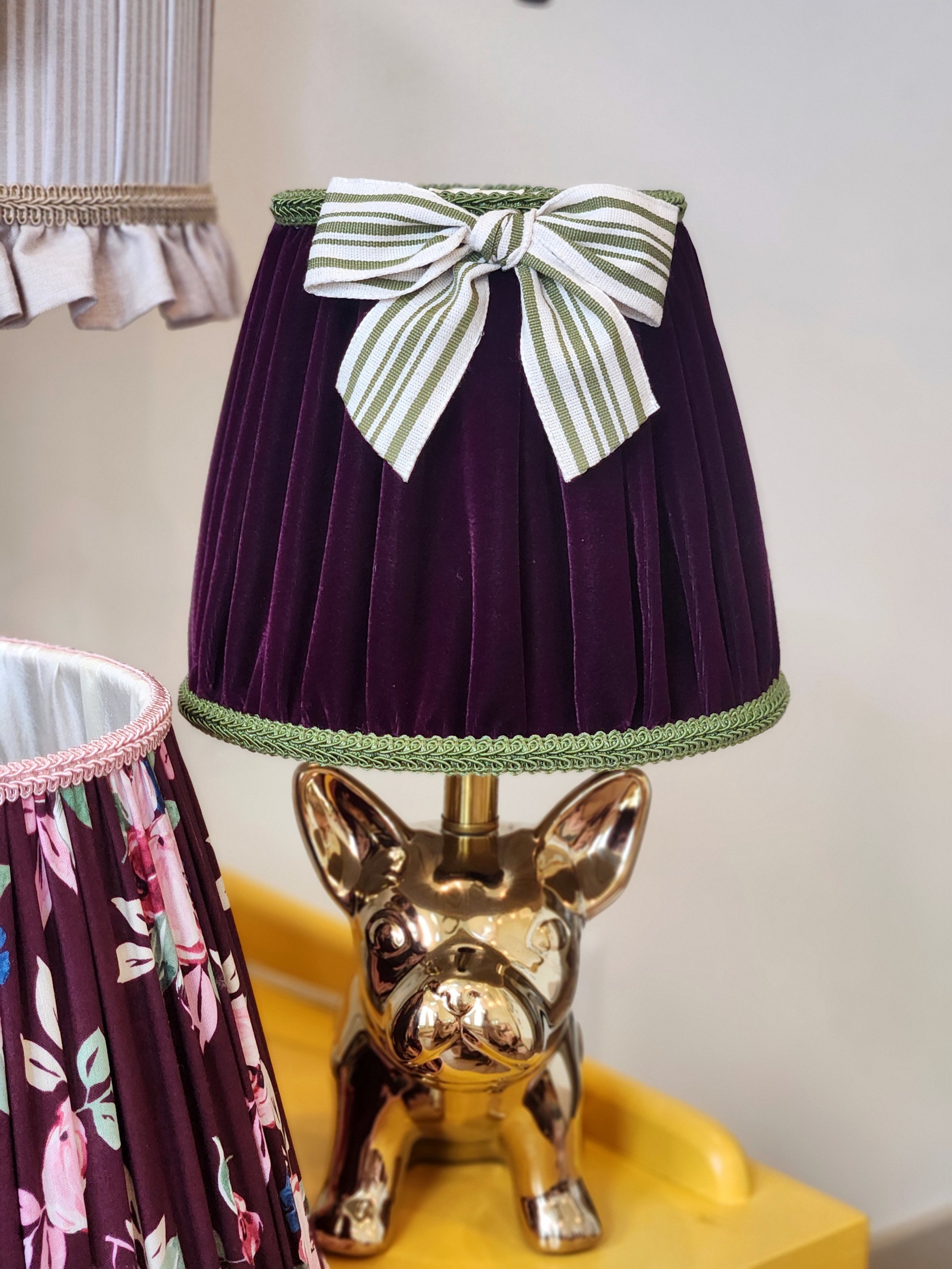 A decorative table lamp with a gold-finished French bulldog-shaped ceramic base and a purple pleated lampshade with a green and white striped bow on top.