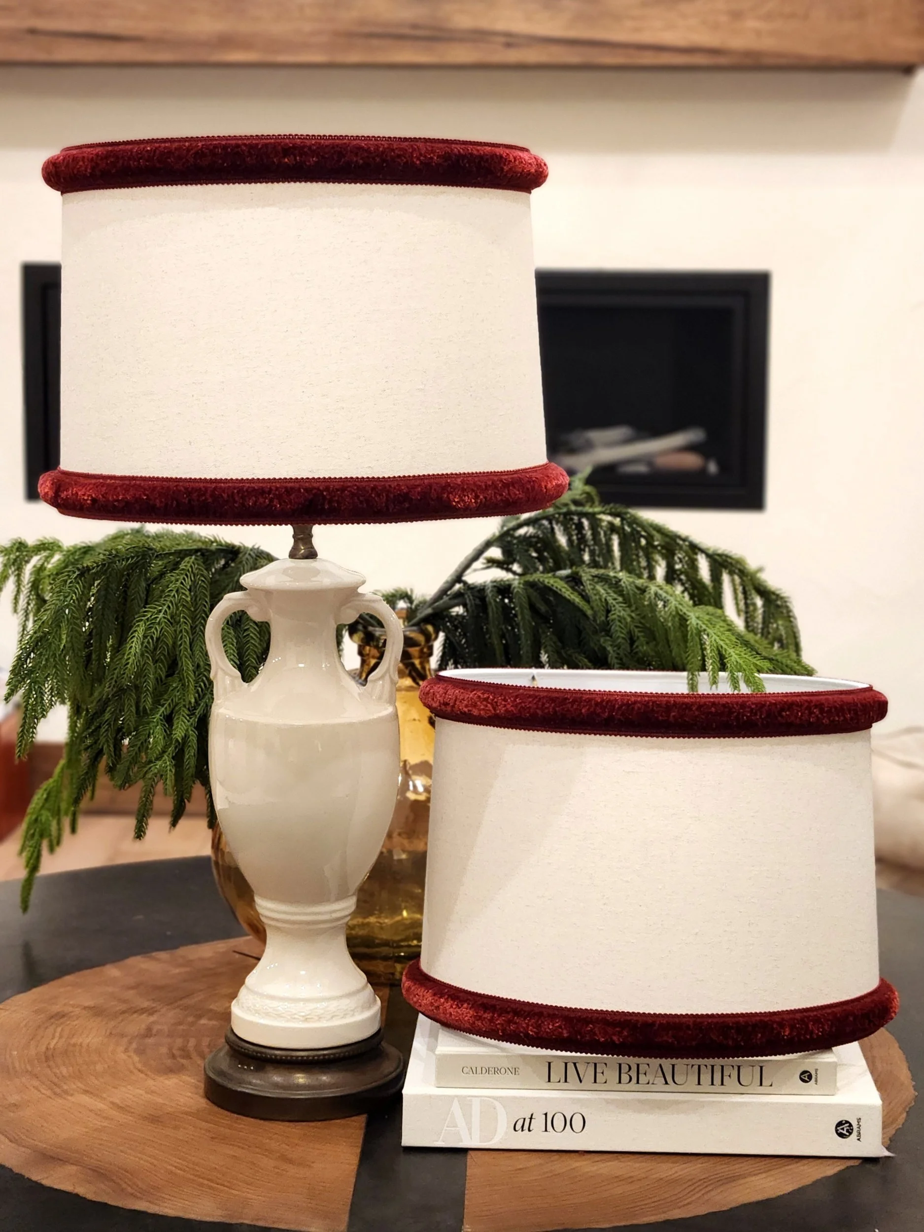 Decorative lampshades, one standing on a ceramic vase and another lying on several books, with greenery and fireplace in background.