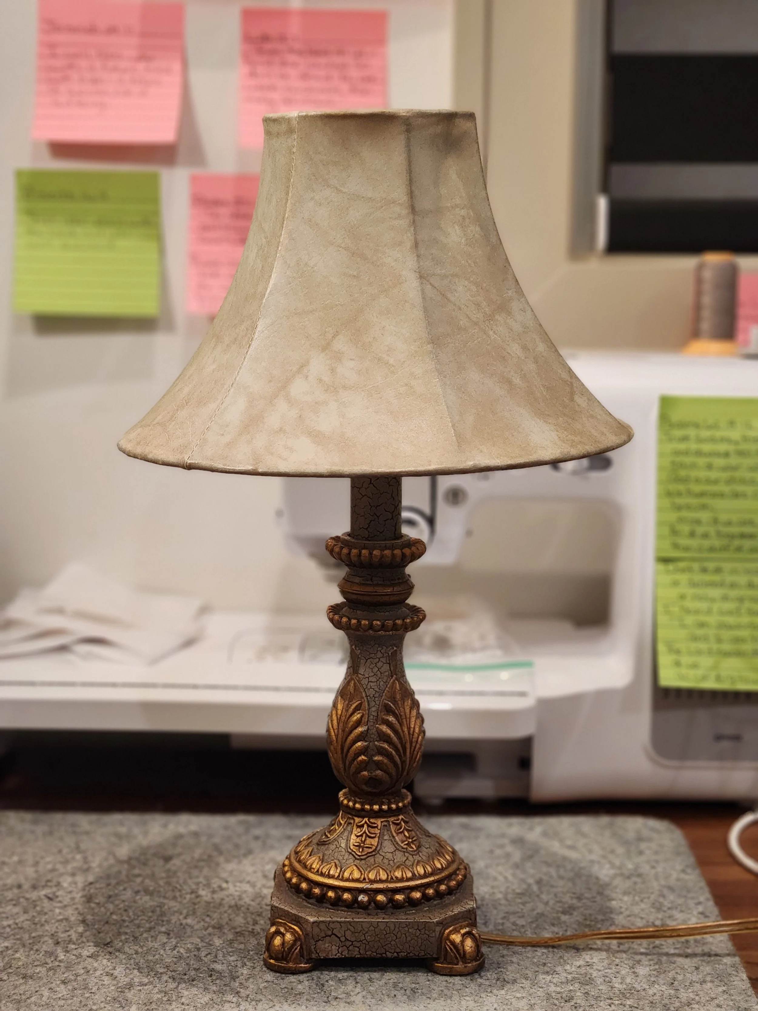 A vintage table lamp with a beige, lampshade and ornate gold and brown base, placed on a gray surface.
