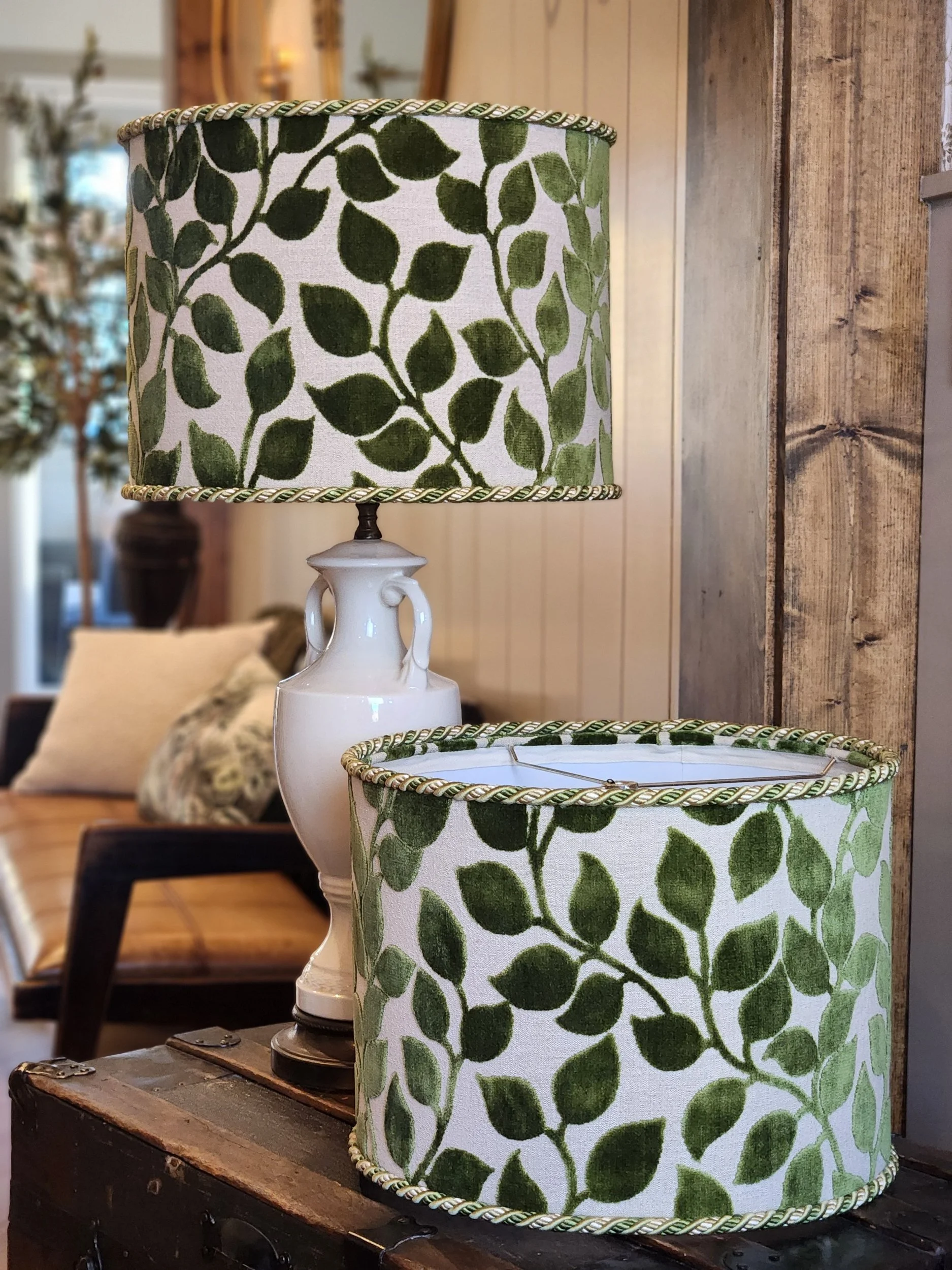 A table lamp with a white ceramic base and a fabric lampshade featuring a green leaf pattern, situated on a wooden surface in a cozy room.