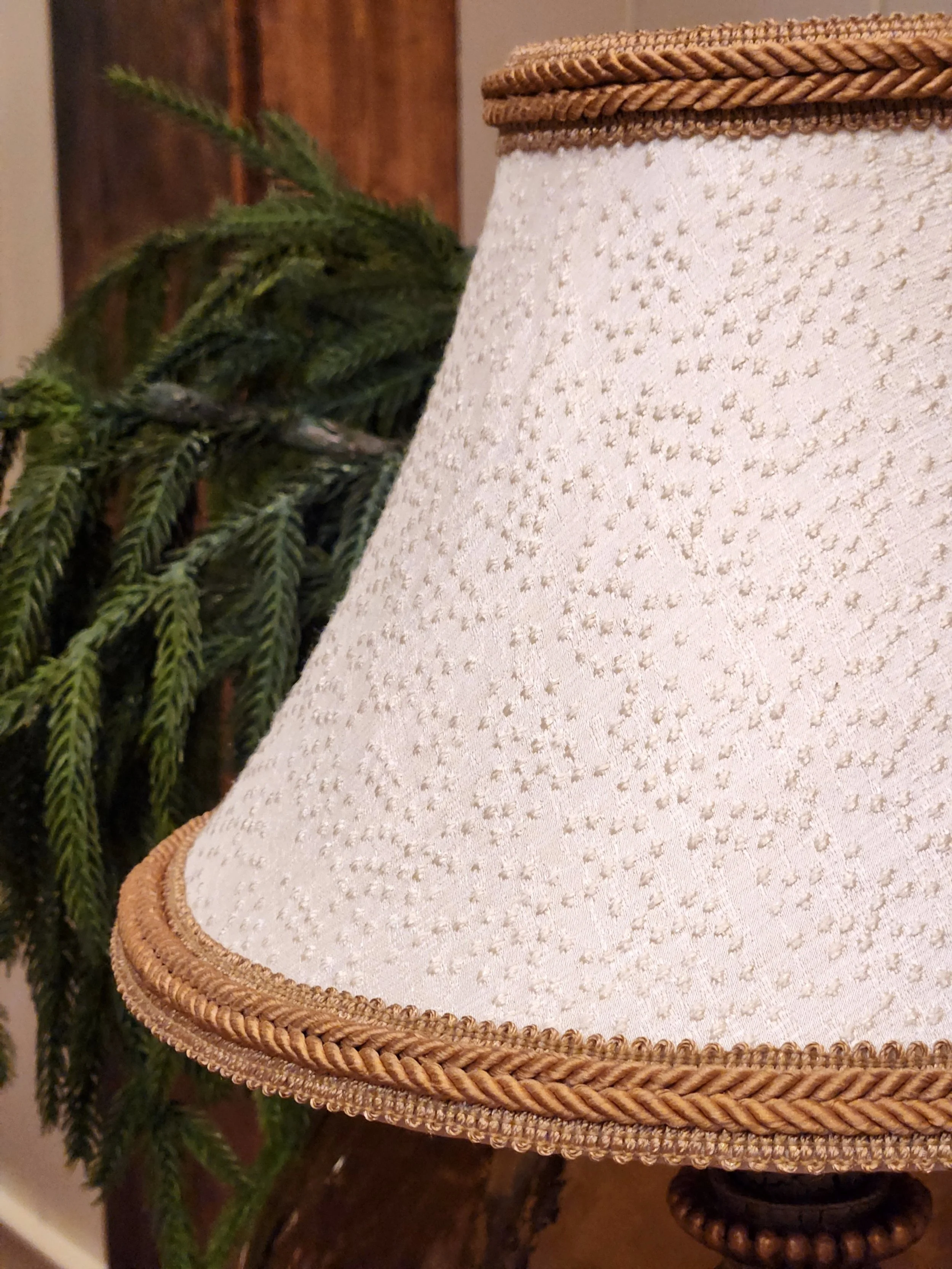 Close-up of a cream-colored lampshade with gold details, next to a green pine branch and a wooden surface background.