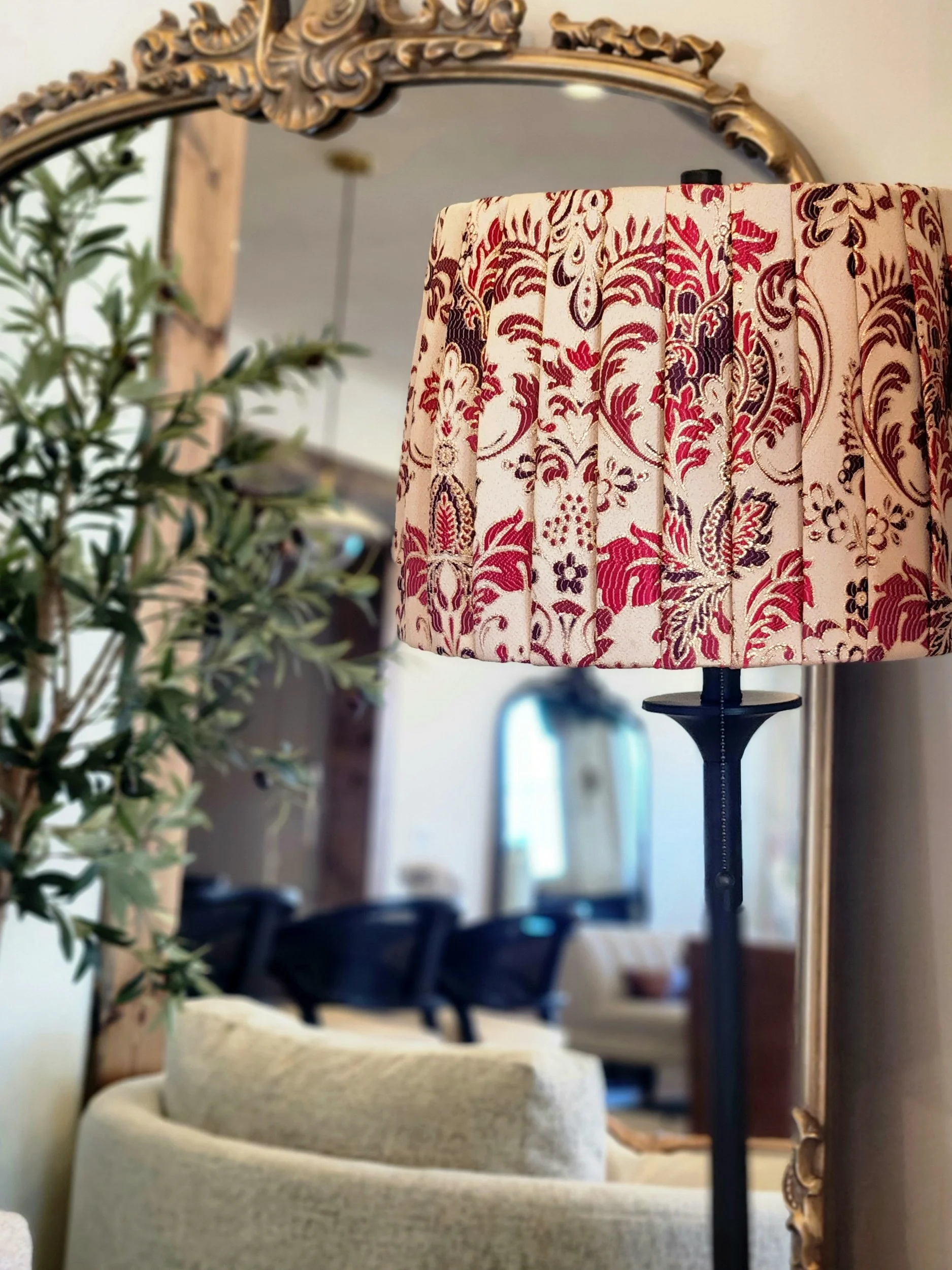 Elegant room decor with ornate mirror and a patterned lampshade on a stand, surrounded by indoor plants and furniture.
