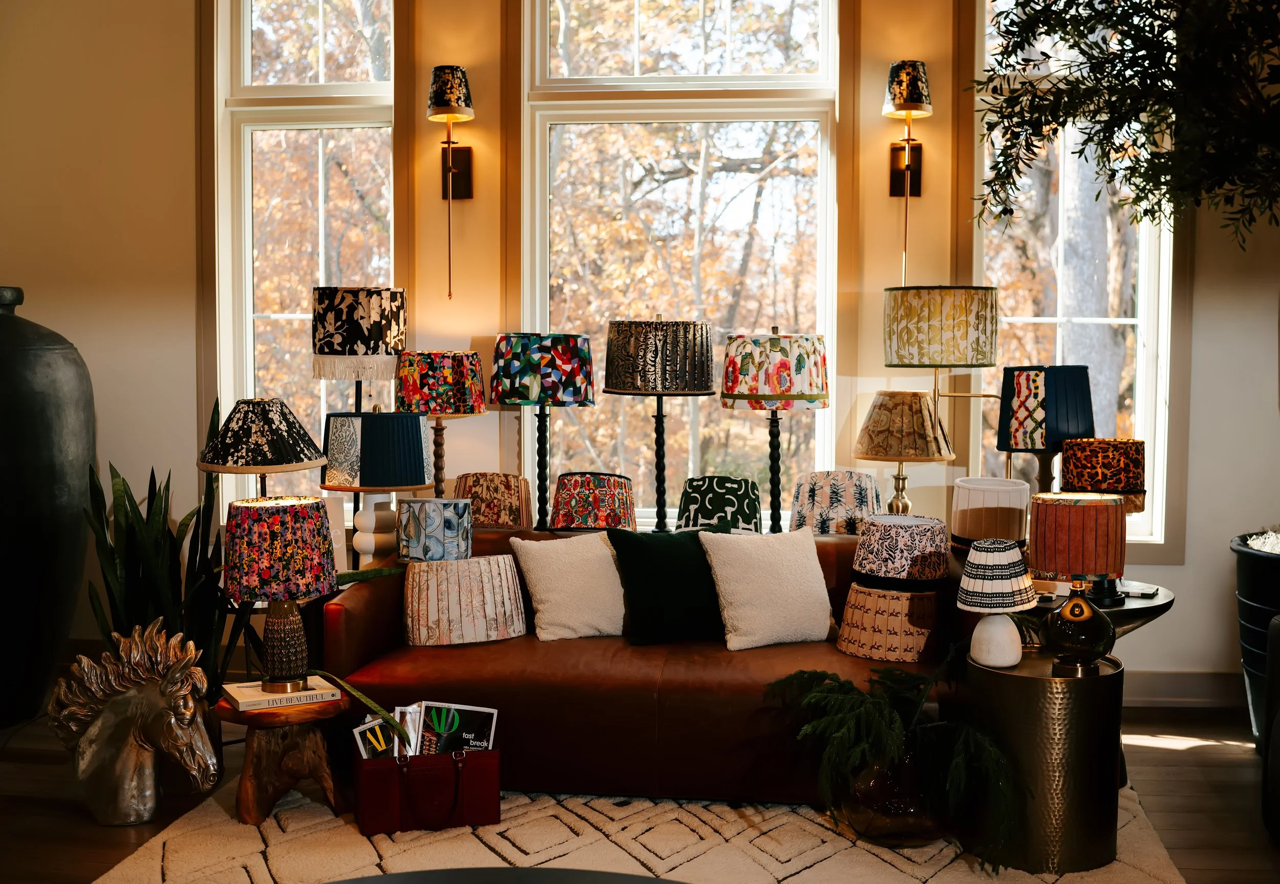 Living room display with multiple patterned lampshades on a brown sofa, decorative pillows, houseplants, and a large window background.