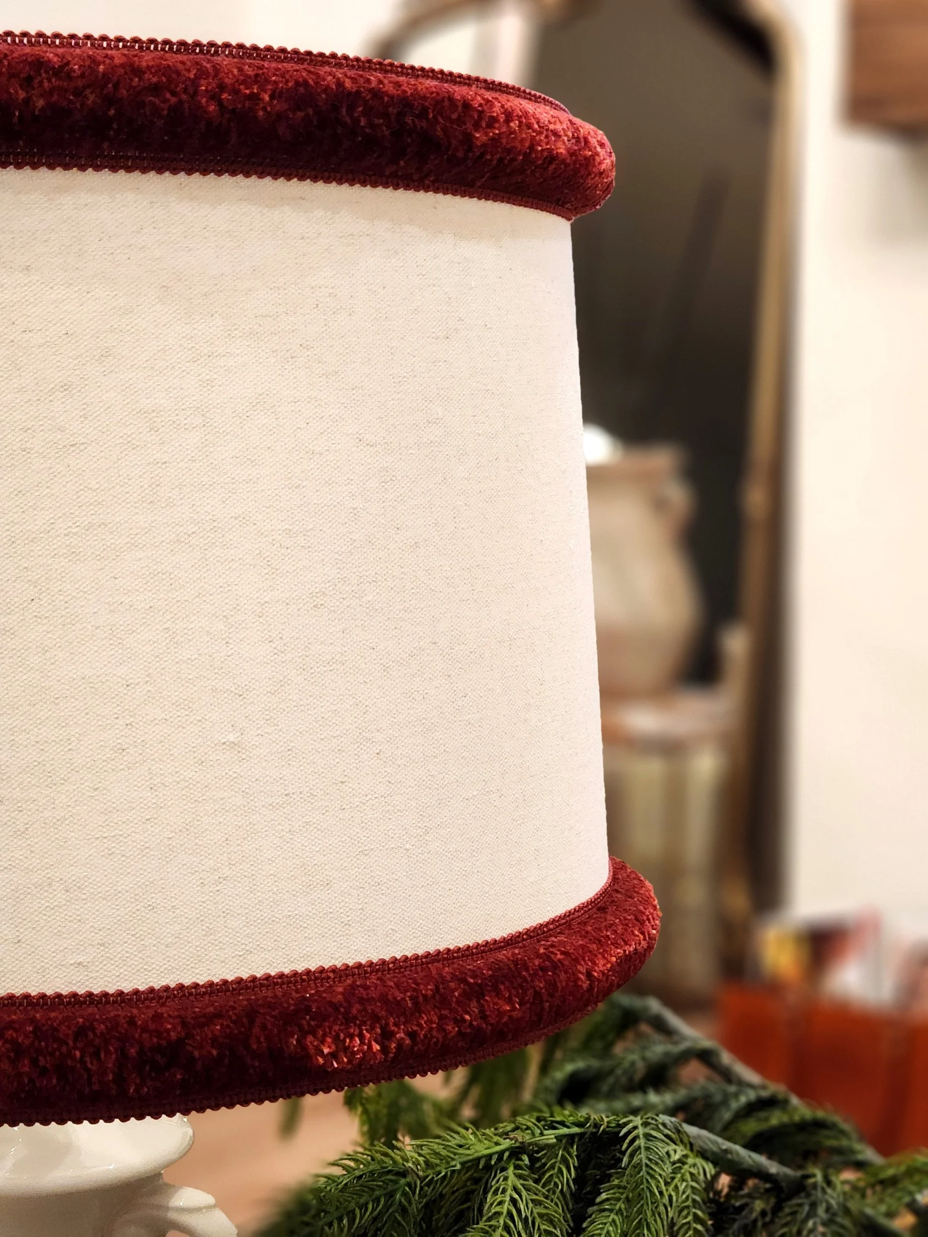 Close-up of a lampshade with a red velvet trim at the top and bottom, placed on a table with some greenery in front.
