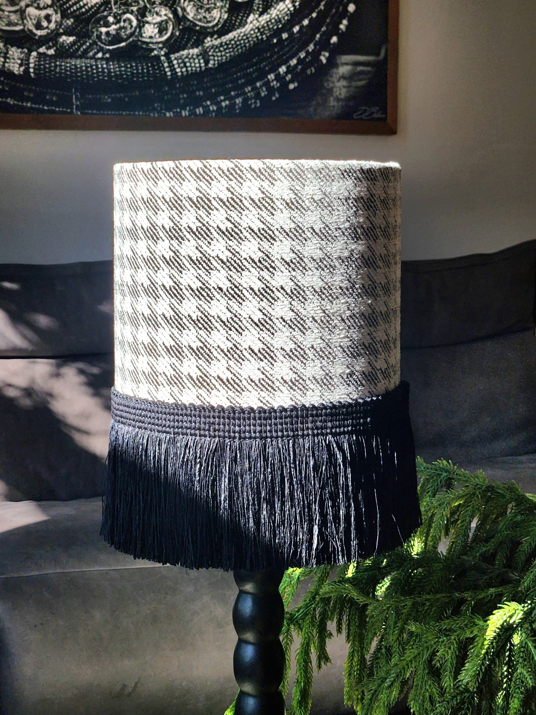 A table lamp with a fringed lampshade, featuring a houndstooth pattern in black and white, next to a green plant on a dark surface.