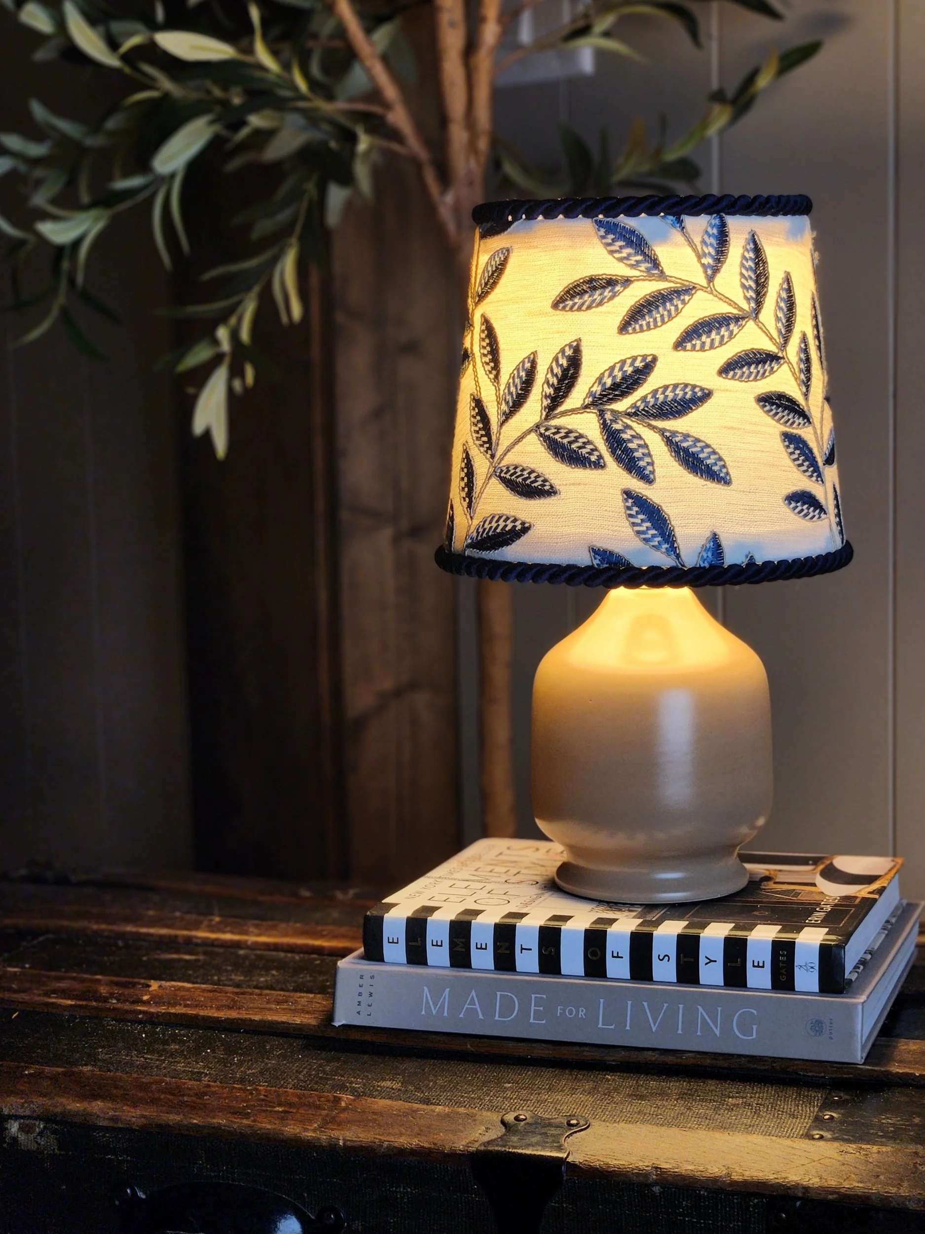 A table lamp with a floral pattern lampshade sits on top of stacked books on a dark wooden surface, with a plant in the background.
