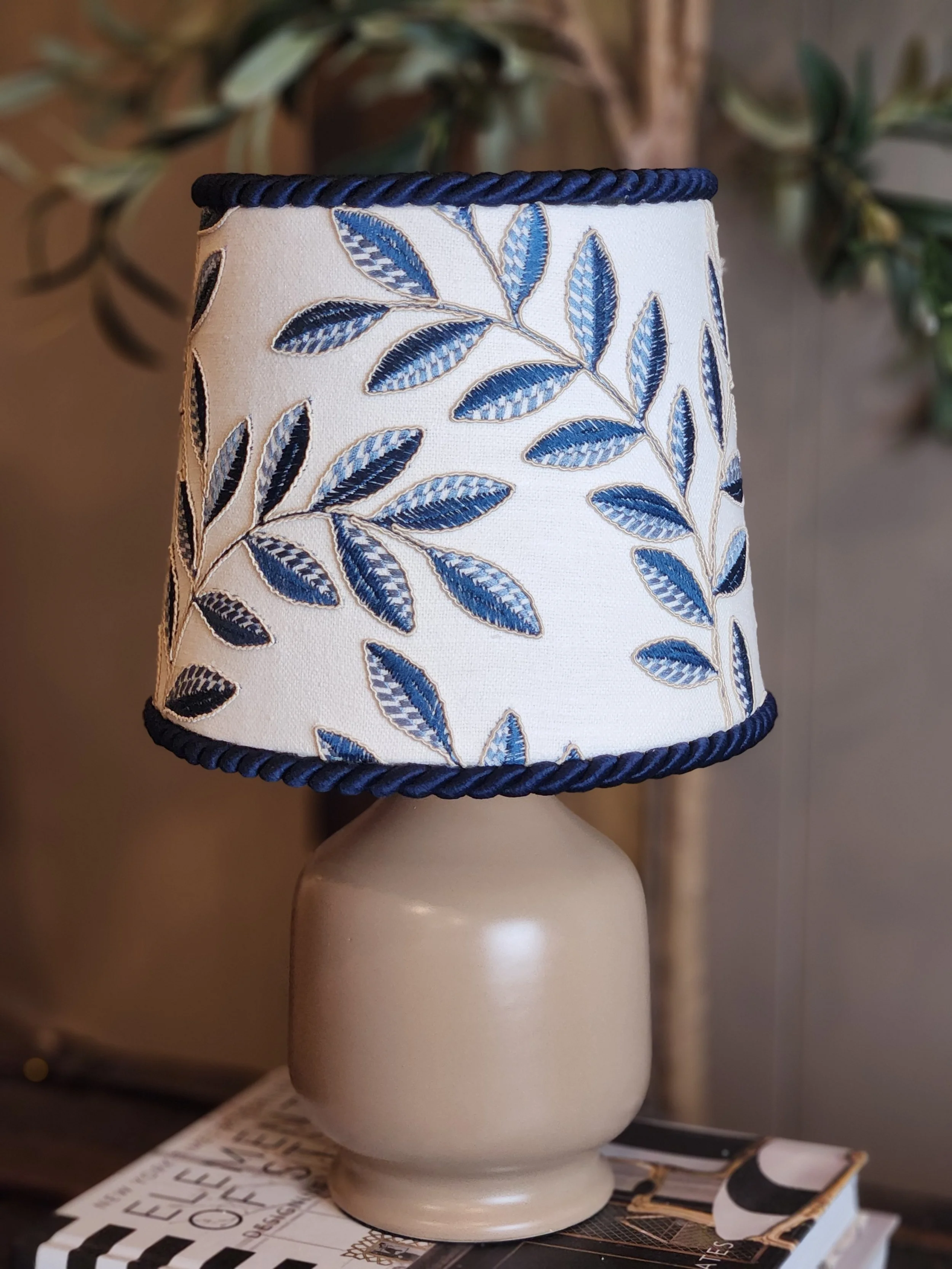 Table lamp with a beige base and a fabric lampshade featuring a blue leaf pattern, placed on top of a magazine.