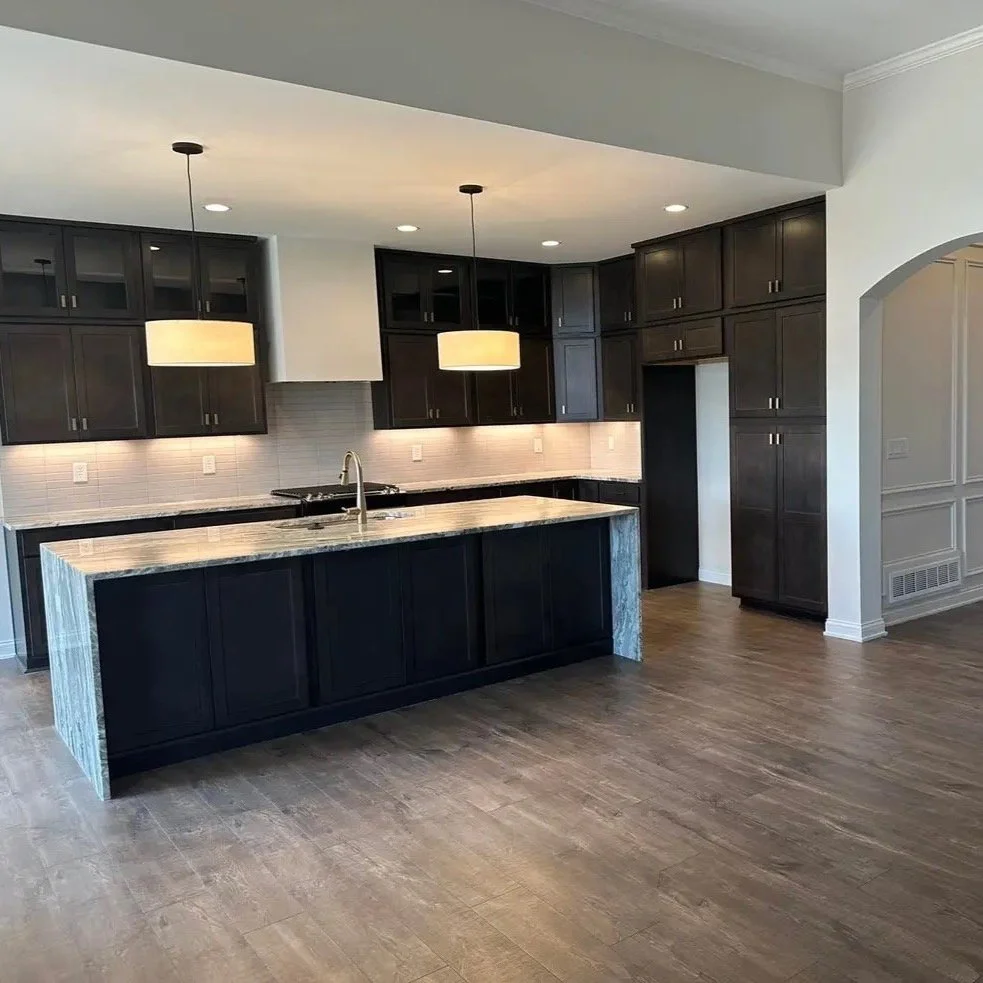 Modern kitchen with dark cabinets and a large island with a marble countertop. Two pendant lights hang above the island, and there are recessed ceiling lights. The kitchen has hardwood flooring and a light-colored tiled backsplash.