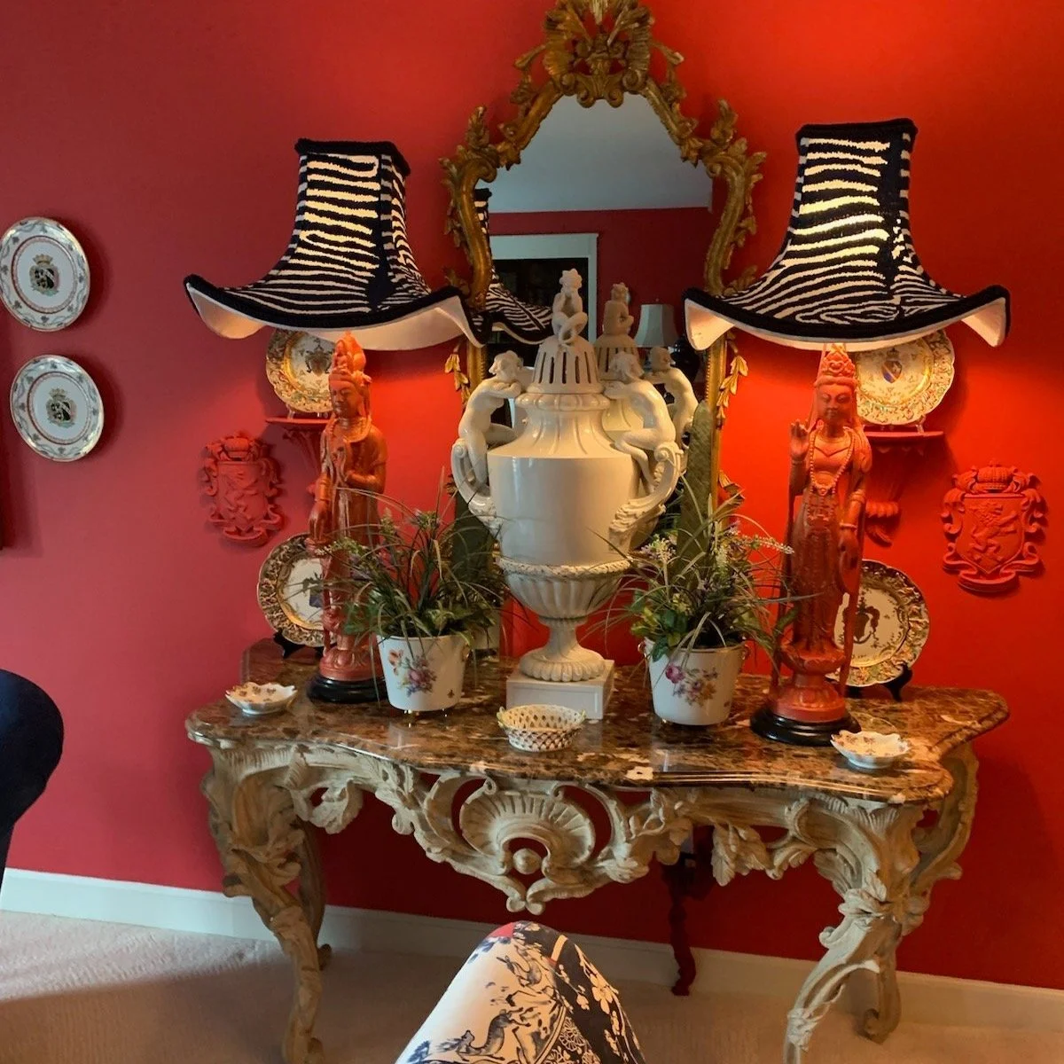 A vintage ornate wooden console table with a marble top, decorated with plants, decorative plates, and figurines, against a red wall adorned with decorative plates, wall plaques, and a gold-framed mirror with two zebra-patterned lampshades.