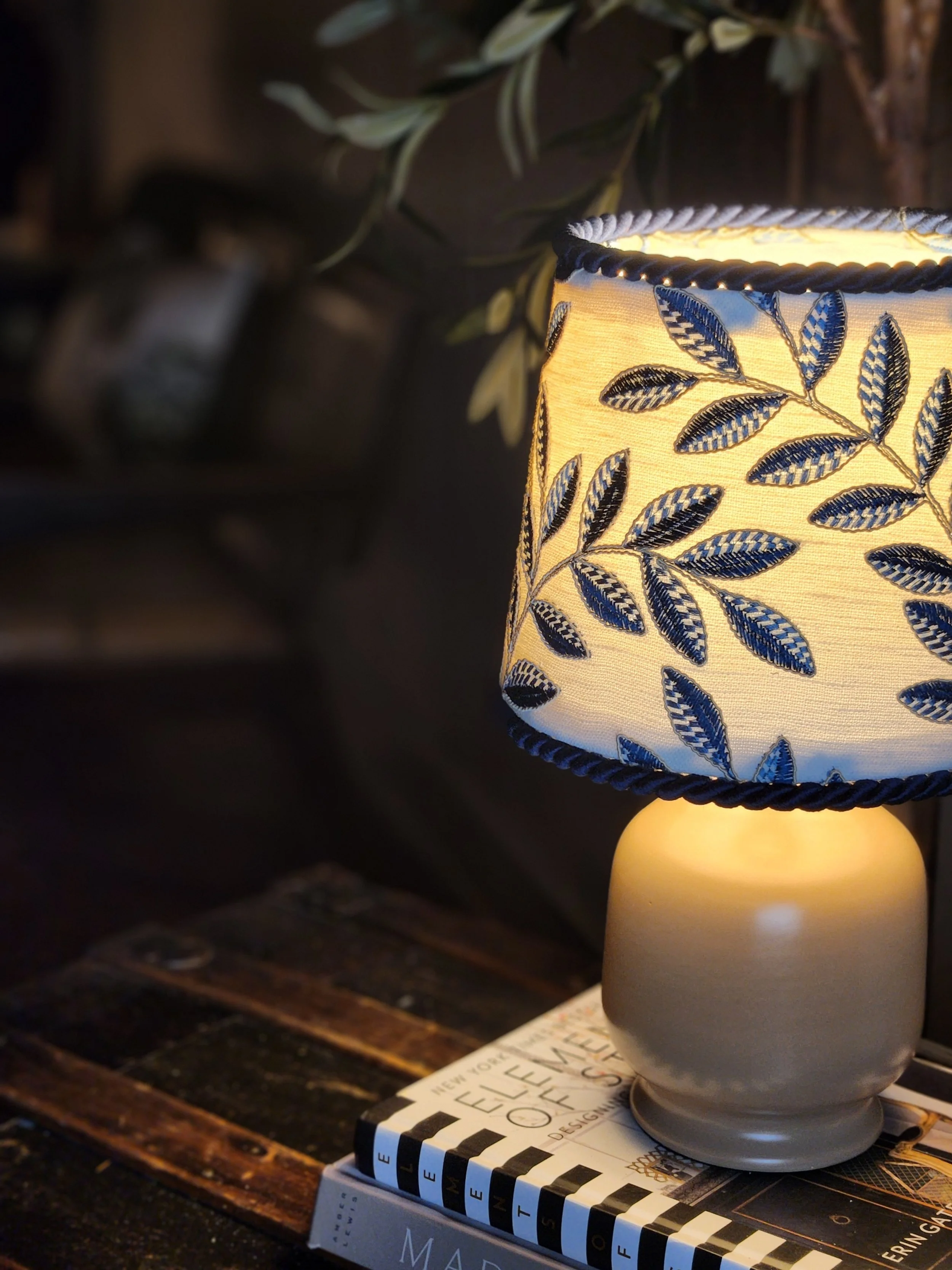 A table lamp with a fabric shade decorated with blue embroidered leaves, placed on top of a stack of design books on a dark wooden table.