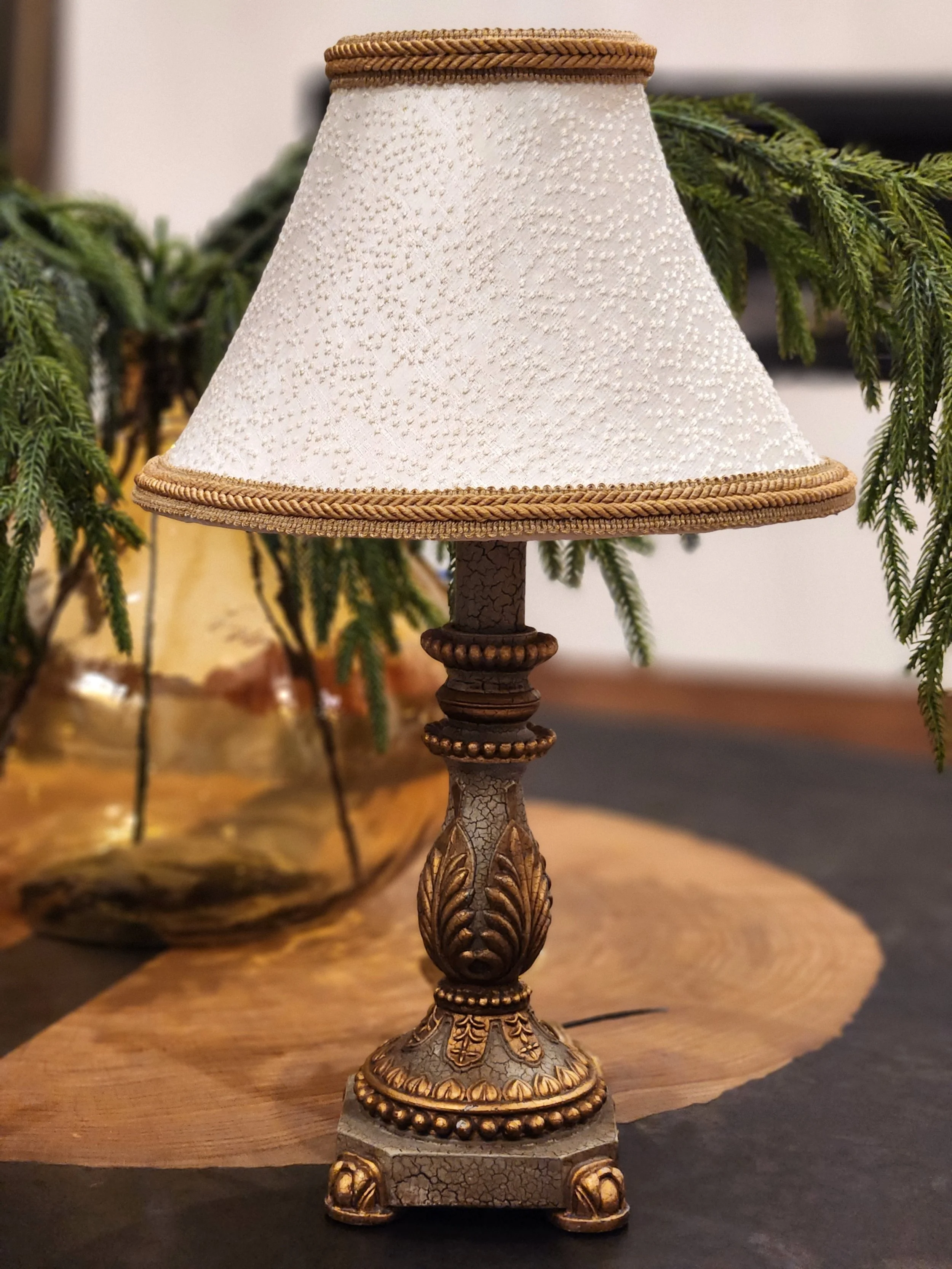 A decorative table lamp with a carved base and a cream-colored lampshade, placed on a wooden surface with greenery in the background.