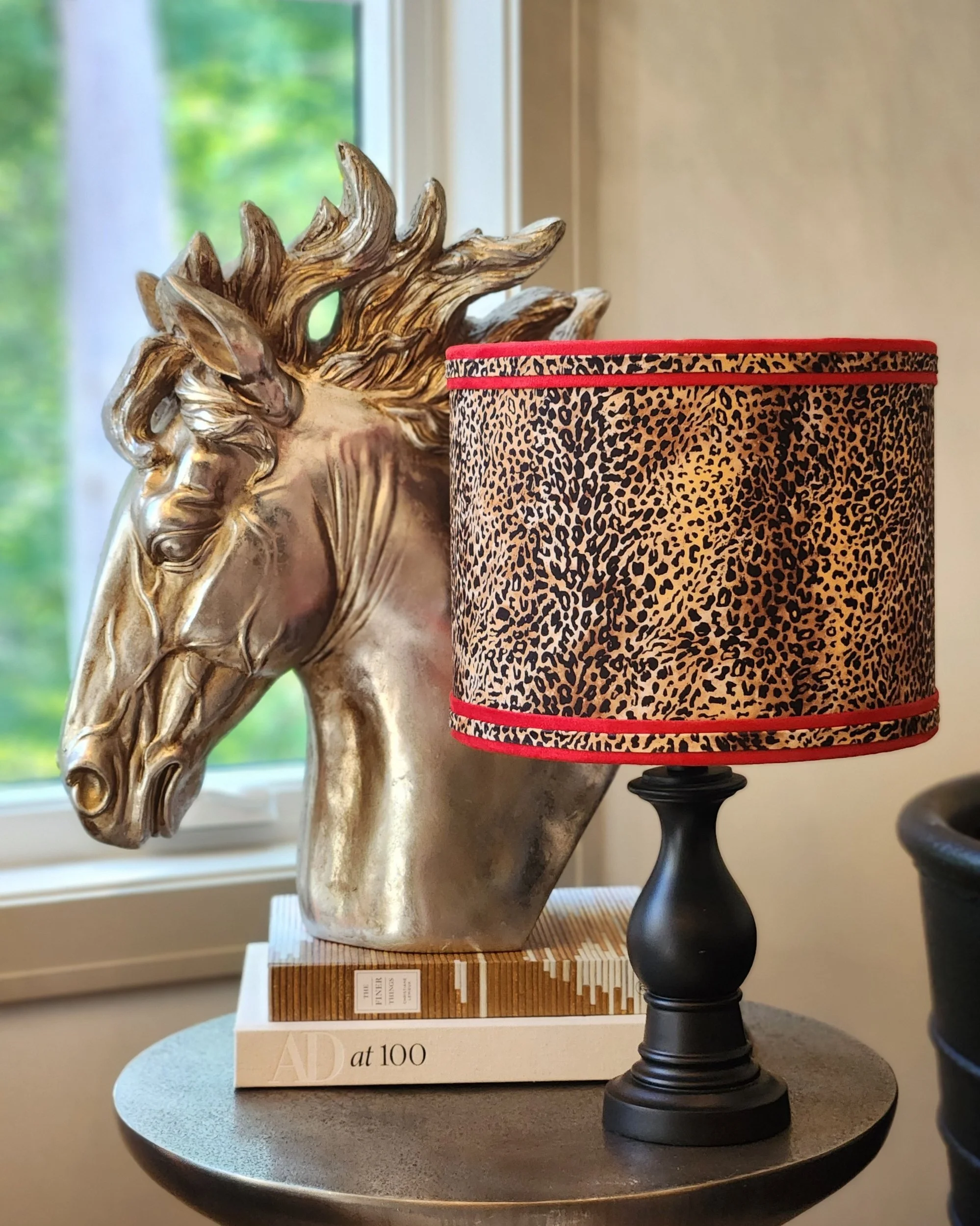 Decorative gold horse head sculpture on books, with a leopard print lampshade on a black stand lamp beside it, placed on a round black table near a window.