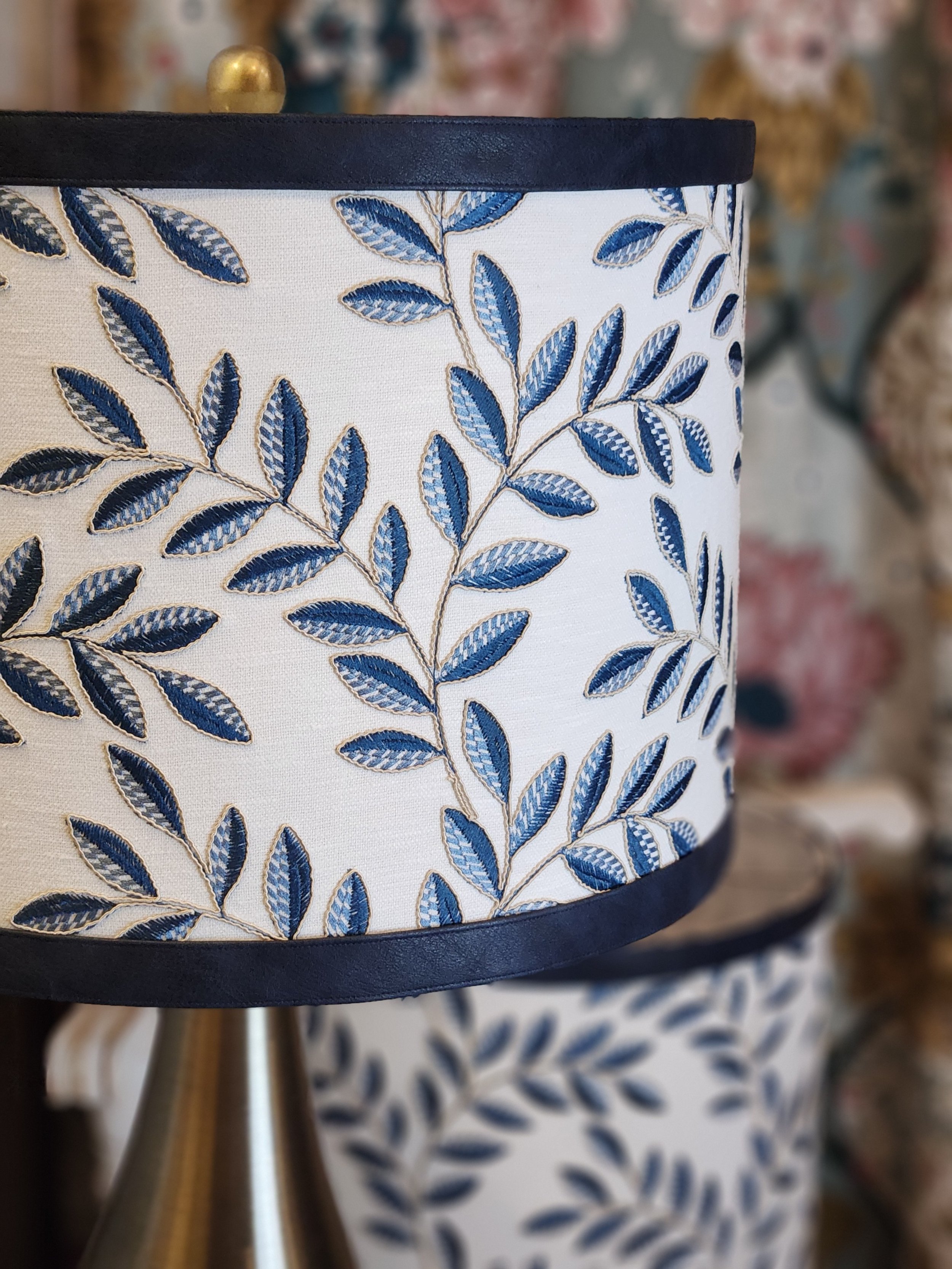 Close-up of a lampshade with embroidered blue leaves on a white background, with a dark trim at the top and bottom.