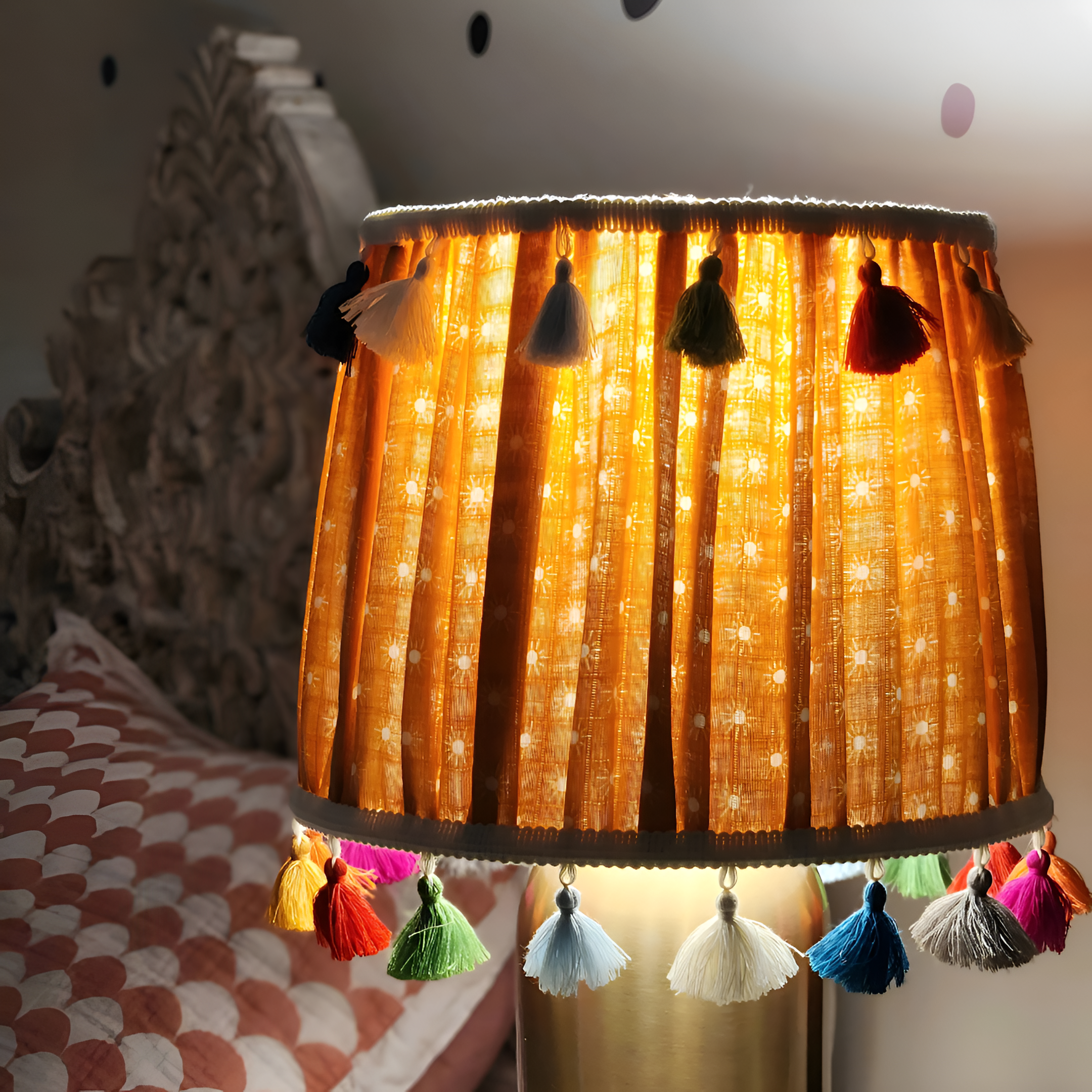 Close-up of a lampshade with a warm orange glow, decorated with colorful tassels hanging around the edge, in a bedroom setting.
