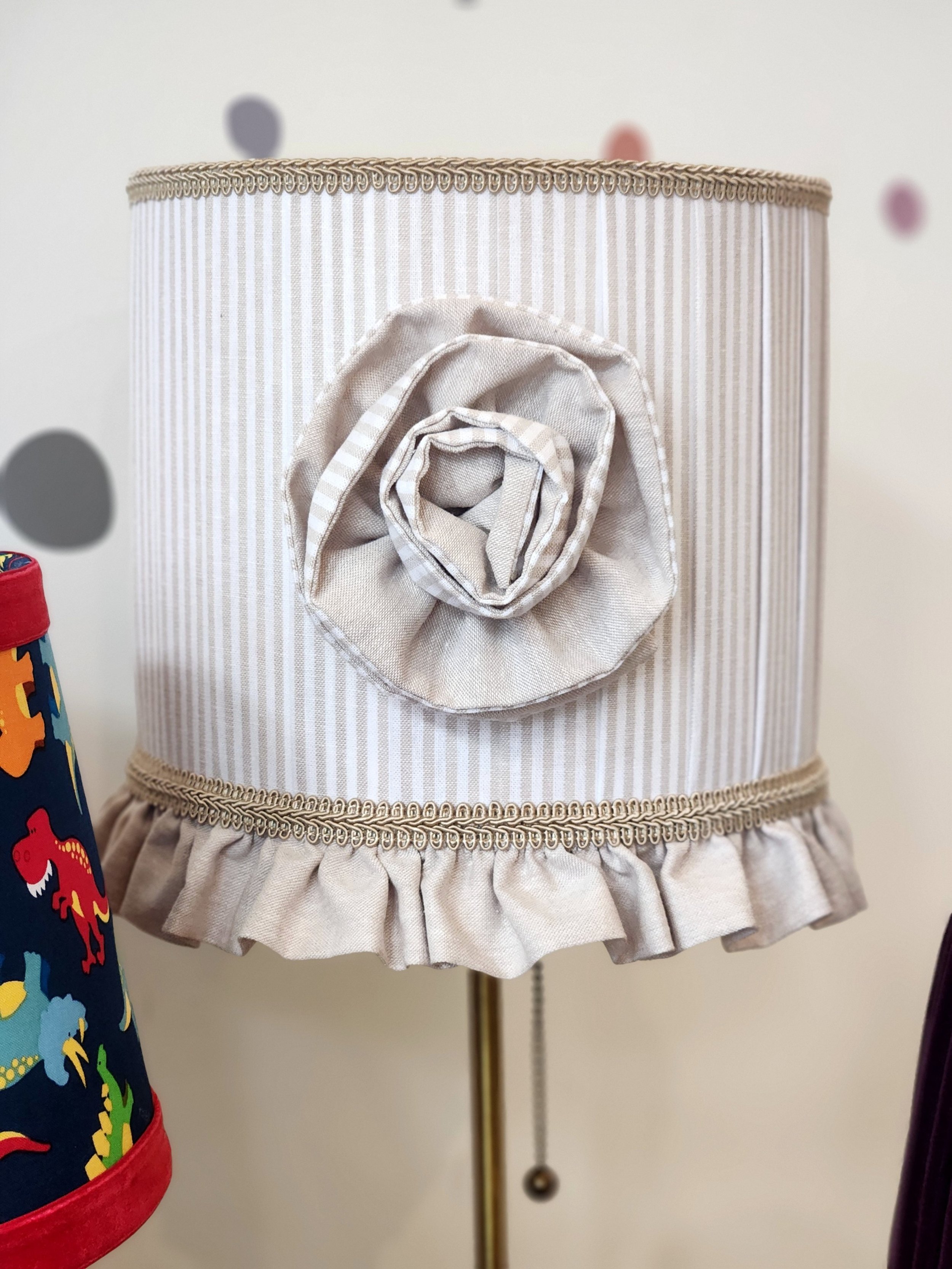 A beige lampshade with a striped fabric covering, adorned with a fabric flower decoration and a ruffled trim at the bottom.