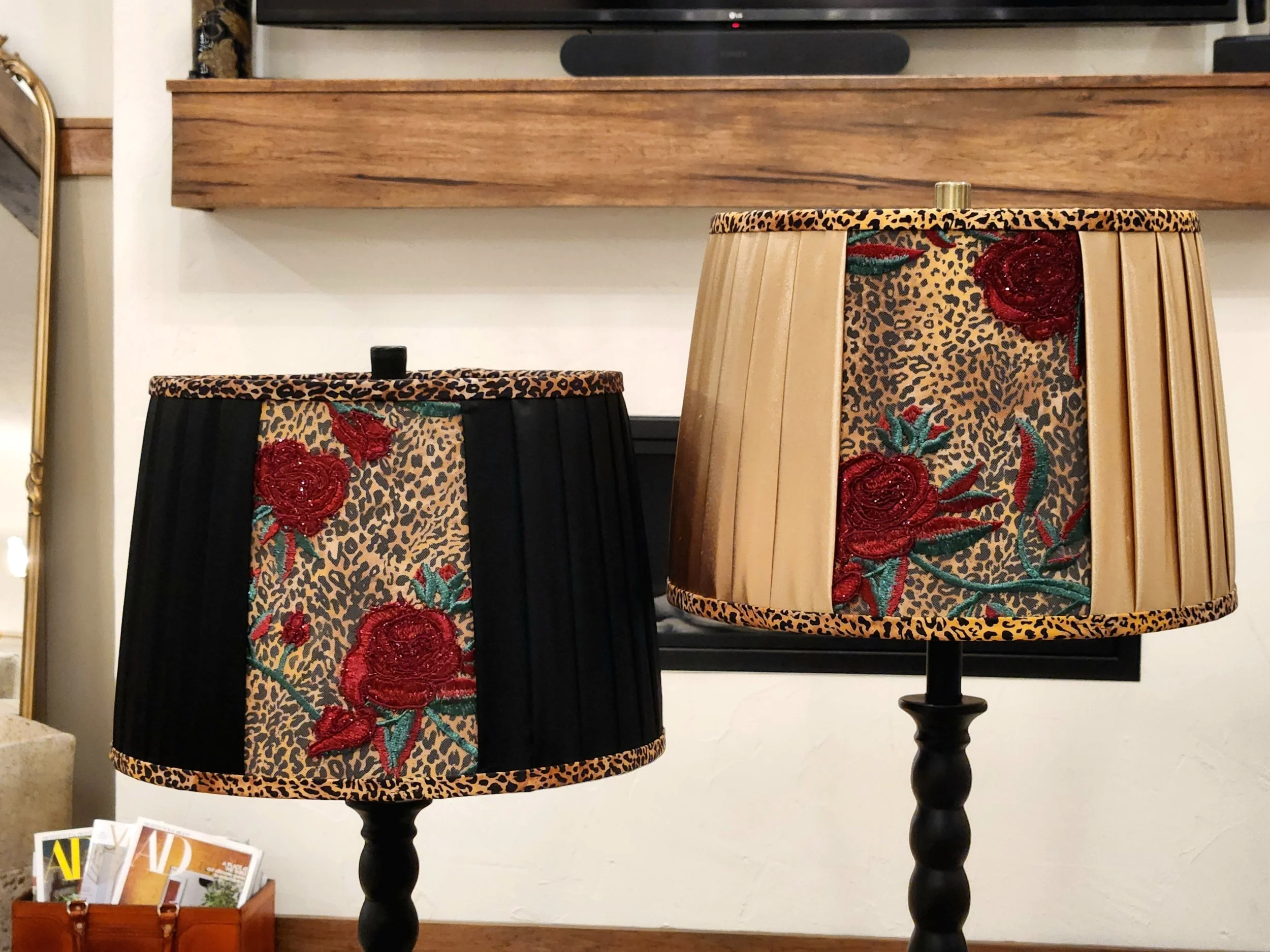Two decorative lamp shades with red rose and leopard print designs, one with black pleats and the other with beige pleats, on dark lamp stands, situated indoors.