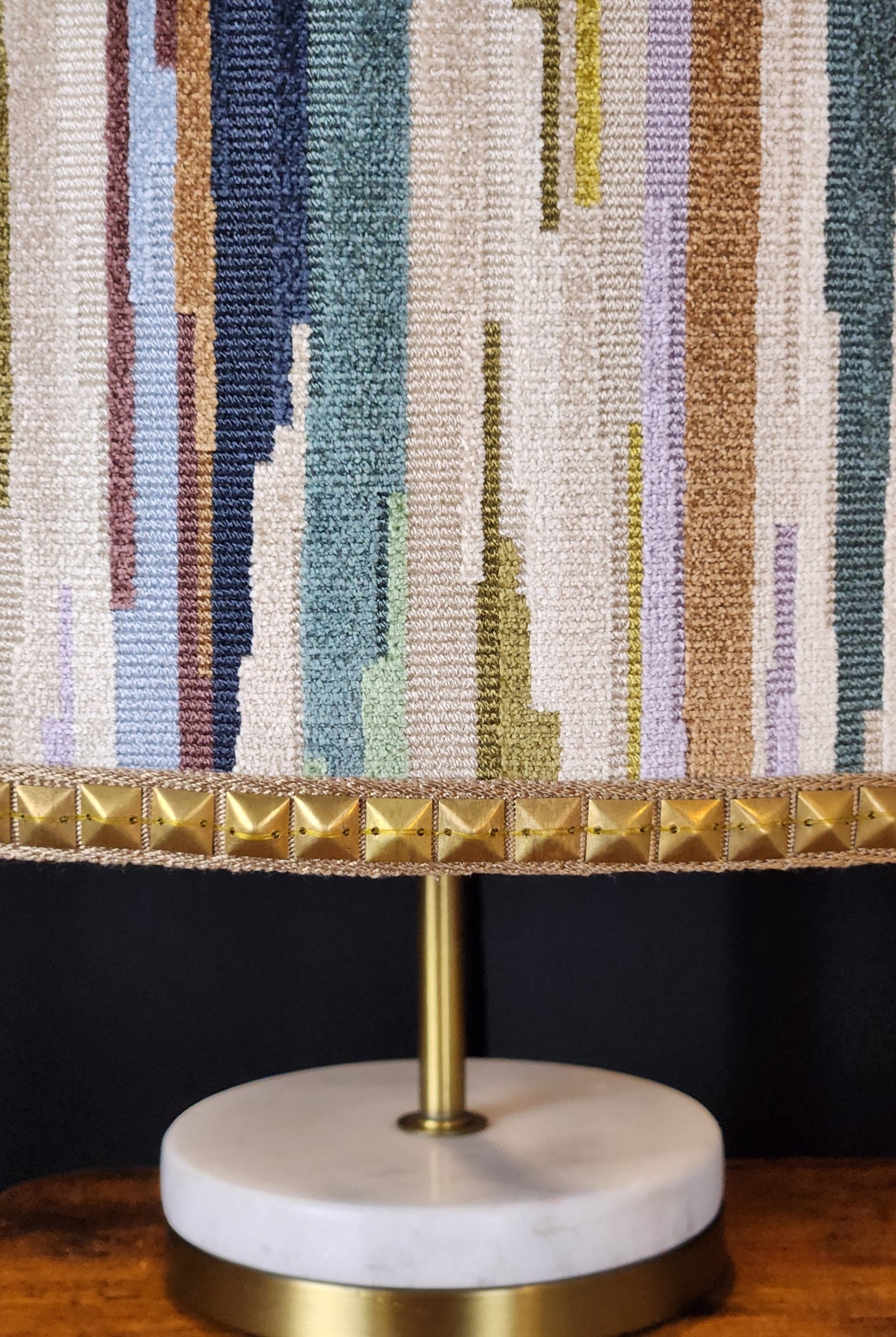 A decorative table lamp with a gold base, white marble bottom, a gold pole, and a colorful striped fabric lampshade. The lampshade has vertical stripes in various colors and patterns, and a row of gold pyramid-shaped embellishments at the bottom edge