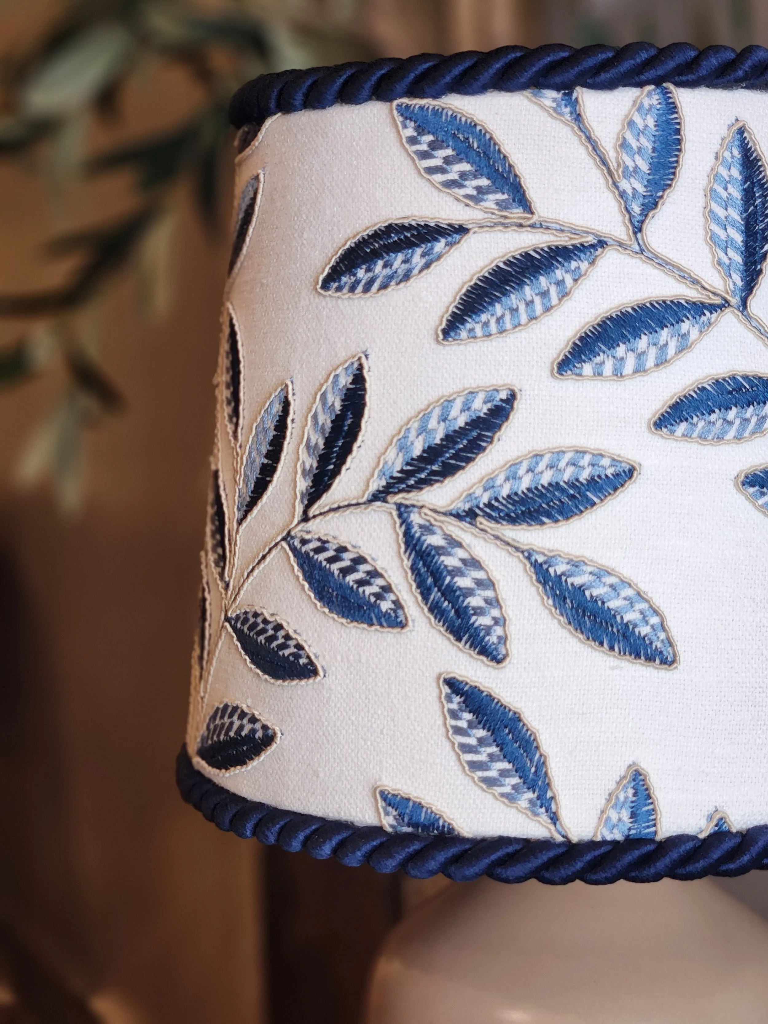 Close-up of a lampshade with embroidered blue leaves on a cream fabric, edged with a navy rope trim.