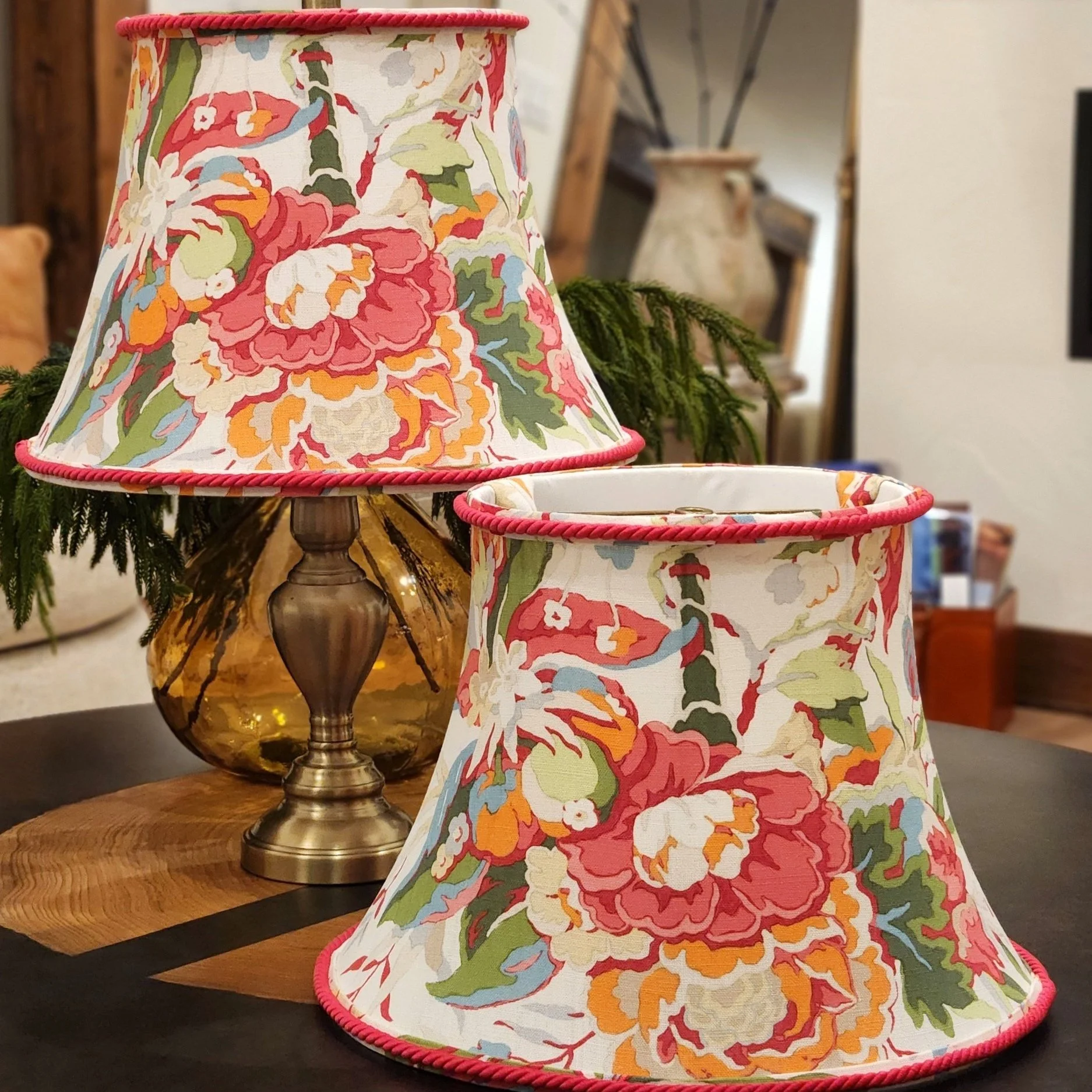 Two lamps with floral fabric shades featuring pink, orange, green, white, and blue flowers, placed on a wooden table near a brass base lamp with a glass body.